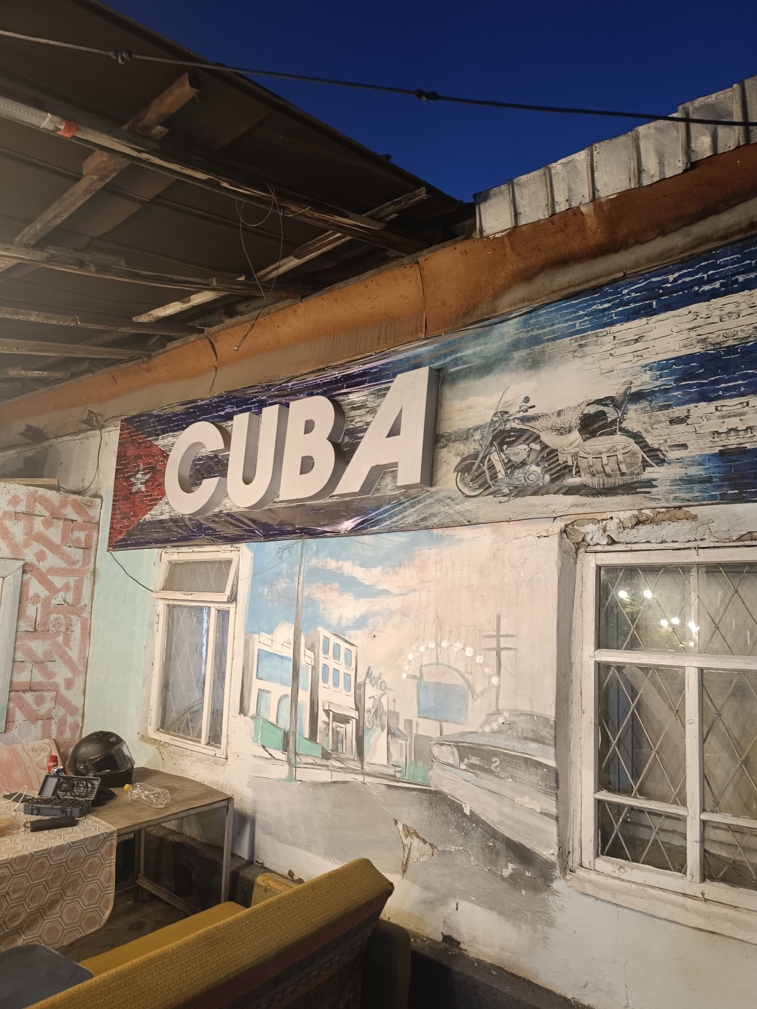 The image depicts a weathered exterior wall adorned with a mural and signage. The mural features a stylized representation of Cuba, with the word "CUBA" emblazoned in large, white, three-dimensional letters. To the left of the text, the Cuban flag is painted, featuring a red triangle and white stripes. To the right of the text, a motorcycle with saddlebags is depicted against a backdrop of blue and white brushstrokes that resemble waves or clouds. Below this, another section of the mural shows a street scene with buildings, a car, and a Ferris wheel, all rendered in a somewhat abstract, faded style.
The wall itself appears to be old and in a state of disrepair, with peeling paint and cracks. Two windows with diamond-shaped panes are visible, one slightly open. To the left of the windows, a table holds a black motorcycle helmet, a small case, and a plastic water bottle. In the foreground, a section of a yellow sofa with patterned cushions is visible. The time of day appears to be dusk or night, as indicated by the dark blue sky and the lighting reflecting in the windows. The overall impression is one of a slightly dilapidated, perhaps outdoor seating area with a strong thematic decoration related to Cuba.