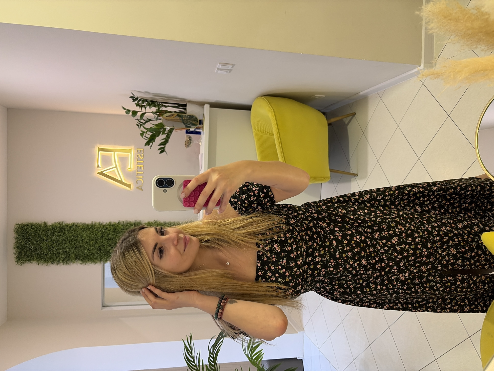 A young woman with blonde hair is taking a selfie indoors. She is wearing a black dress with a pink floral print. She is holding a smartphone with a white case and a pink pop socket. The environment appears to be a stylish interior. There's a yellow chair, a wall with a decorative green panel and a golden lighted sign "FA Estetica" is also visible. Based on the sign, the location is likely "FA Estetica" in Budva, Montenegro.