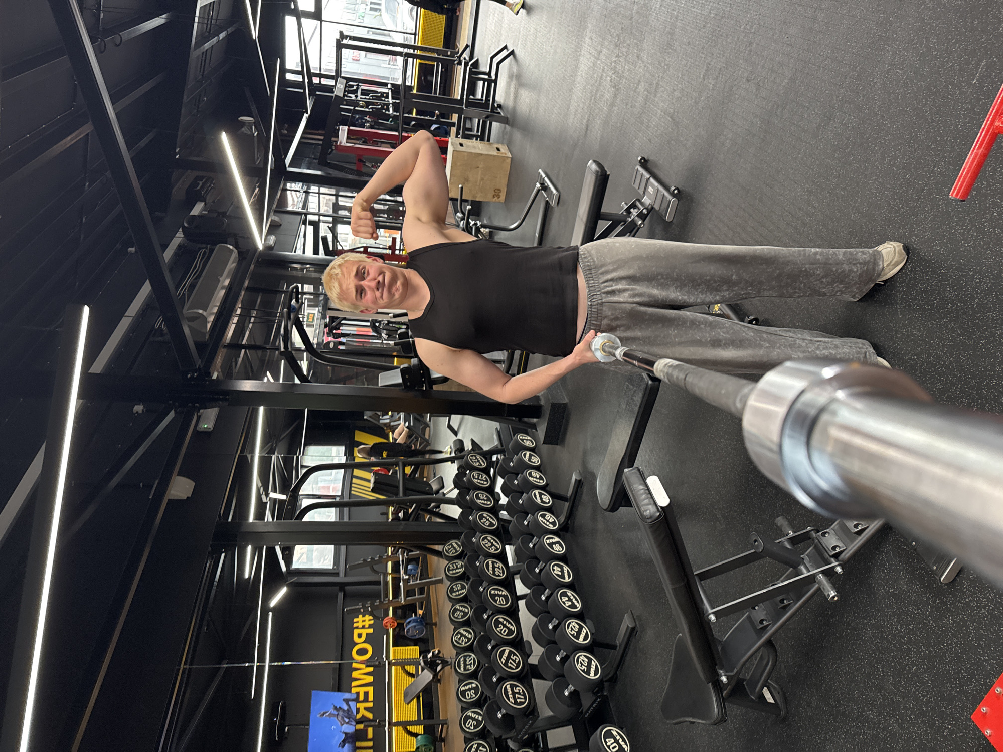 The image, rotated 90 degrees clockwise, captures a man posing in a modern gym. The man, with short blonde hair, is wearing a black sleeveless tank top and grey sweatpants. He is smiling directly at the camera while flexing his right bicep and holding a chrome barbell with his left hand, possibly using it as a selfie stick.
The gym features a dark aesthetic with black walls, a black speckled floor, and prominent linear LED lighting strips on the ceiling and walls, creating a sleek, industrial look. Large windows in the background allow natural light into the space, suggesting it is daytime.
The gym is well-equipped with various fitness gear. To the right of the man is a long rack of dumbbells, neatly organized by weight. Visible text on the dumbbells includes "ZIVA," "AVIS OS," and various weight numbers such as "30," "25," "22.5," "20," "17.5," "46," "44," "42.5," "40," and "52." Other gym equipment visible includes weight benches, several exercise machines, and a wooden plyometric box labeled "30" in the background. A yellow sign or banner on the wall bears the text "#BOMFK." The overall atmosphere suggests a functional and well-maintained fitness facility.
