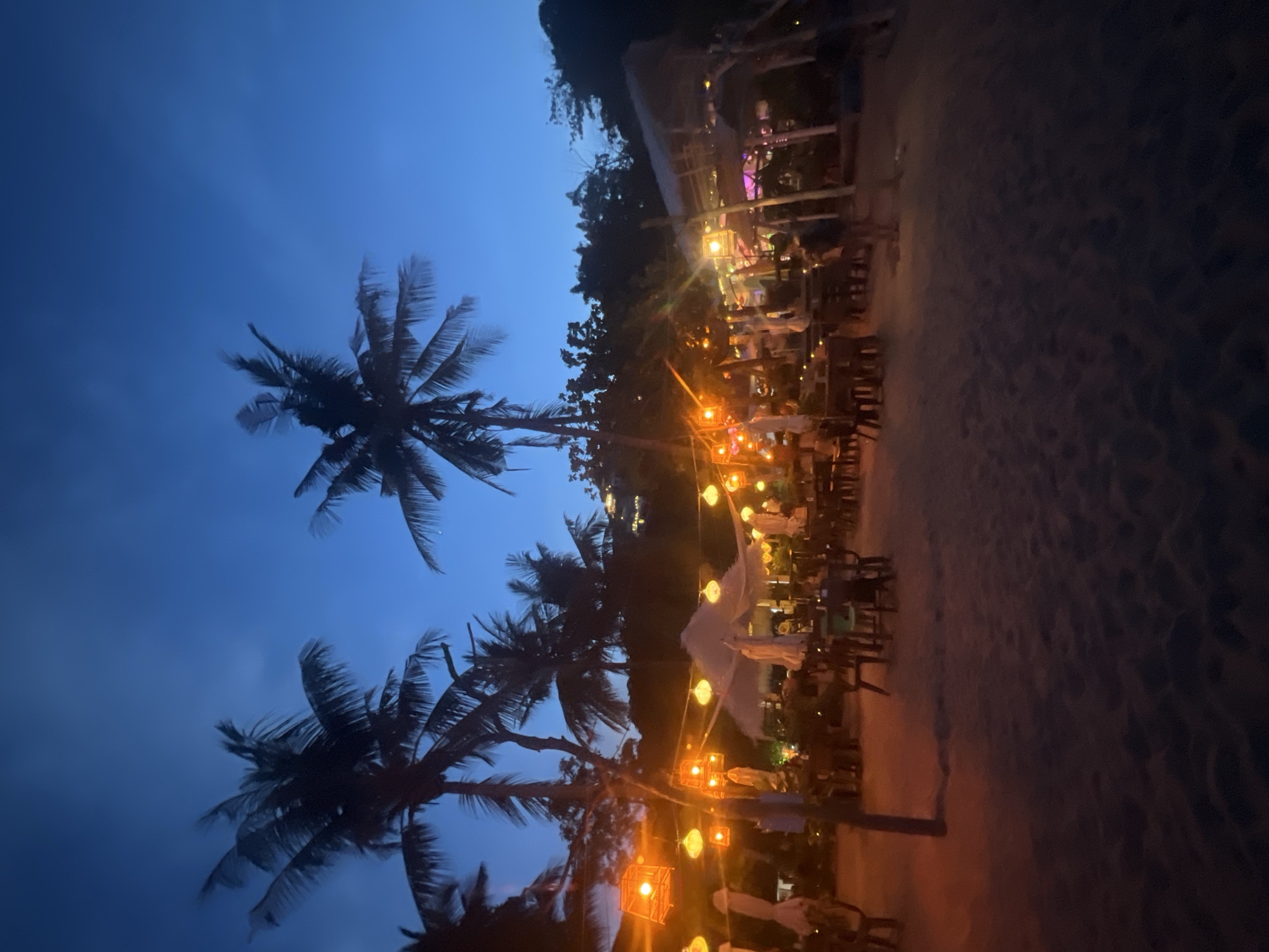 In the city of Ko Samui District, Thailand, at dusk, a beachside restaurant is illuminated by strings of orange paper lanterns. Silhouetted palm trees stand tall against the deep blue twilight sky. Tables and chairs are set up on the sandy beach, some with umbrellas providing shade. The atmosphere is serene and inviting, suggesting a place for evening dining or relaxation.