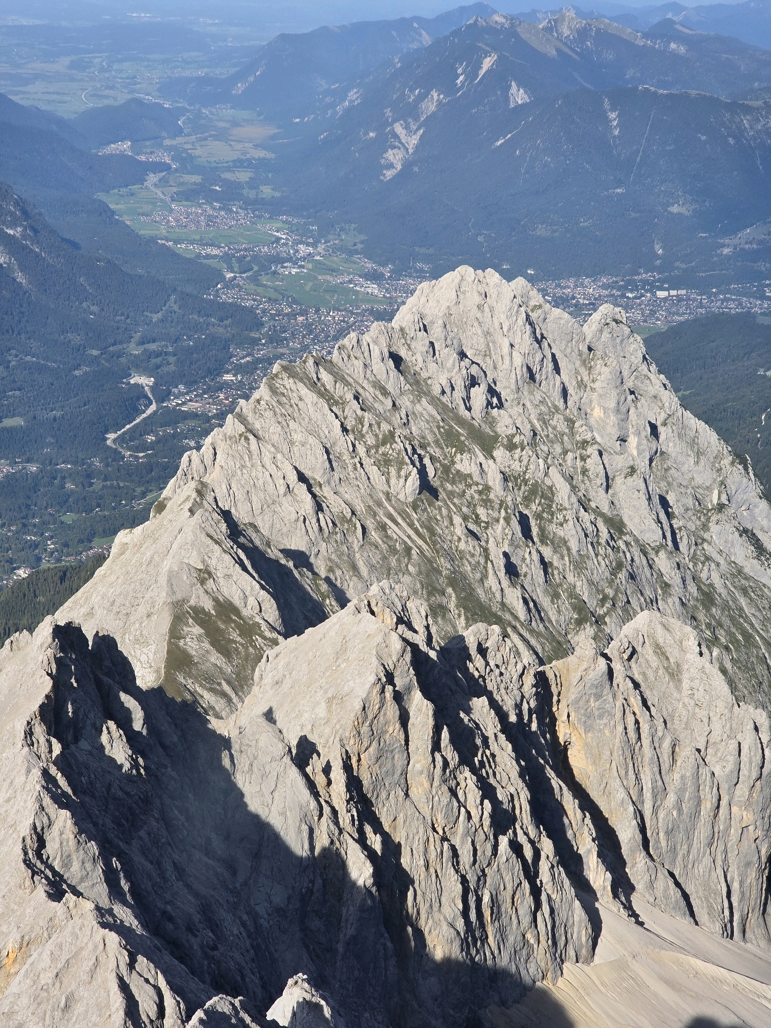 This is a high-angle, full-frame, natural light photograph of a rugged mountain range with a valley and a town visible in the distance. The foreground is dominated by steep, jagged grey rock formations, with patches of green vegetation clinging to some slopes. Deep shadows are cast across the rocky terrain, highlighting its textures and dramatic contours.
In the middle ground, the mountain range continues, showing steep slopes covered in dark green forests. A town, identifiable by its densely packed buildings and church spires, is nestled in the valley. The town appears to be Grainau, Germany, given the context of the provided crops and its characteristic alpine setting. A road or highway is visible winding through the valley, connecting the town to other areas.
The background consists of more distant mountain ranges, fading into a hazy blue sky. The overall scene conveys a sense of vastness and natural beauty, with the human settlement appearing small and integrated within the grand mountain landscape. There are no people visible in the image.