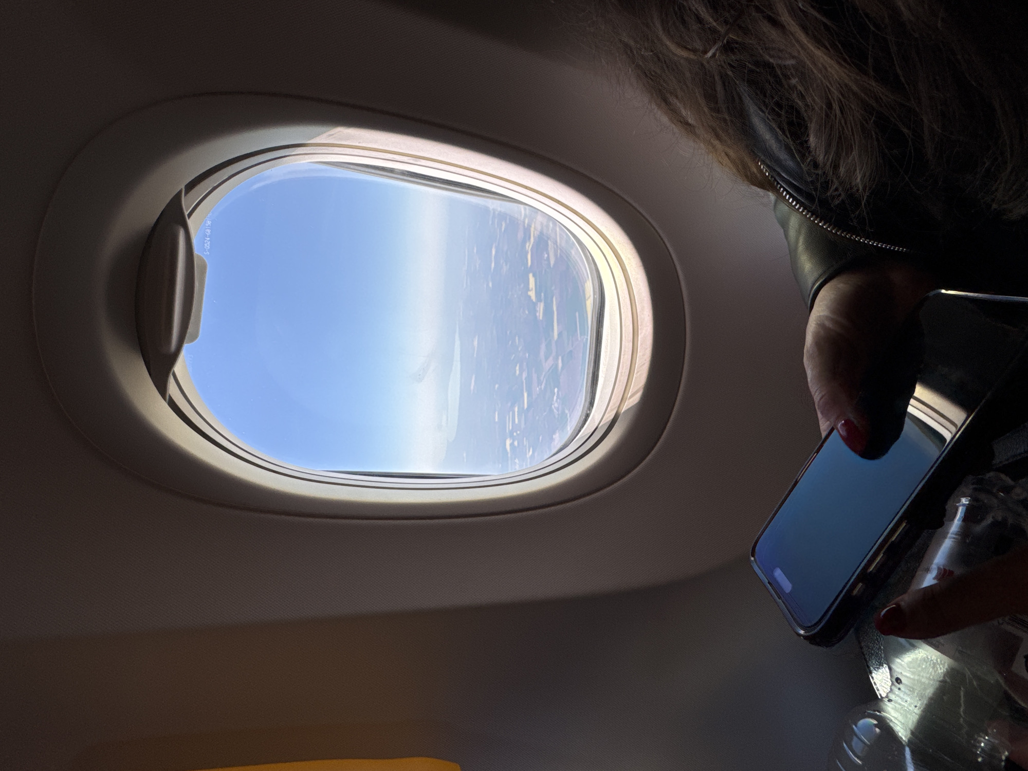 An interior shot from an airplane captures a passenger looking out a window during daylight. The circular window reveals a clear blue sky and the earth below, which appears as a patchwork of fields and possibly water bodies, indicating flight at cruising altitude. A partial view of a person is visible on the right side of the frame; they have dark hair and are wearing a black leather jacket with a metallic zipper. The person's hand, adorned with red nail polish, holds a black smartphone with a reflective screen and a clear plastic bottle. The airplane's interior wall and ceiling are light beige. A window latch mechanism is visible on the left side of the window. Faint text, "S-0324-63(3R)", is visible on the window pane itself. The scene depicts air travel, potentially in the vicinity of Sofia, Bulgaria, under bright, clear weather conditions. No specific emotions are discernible.