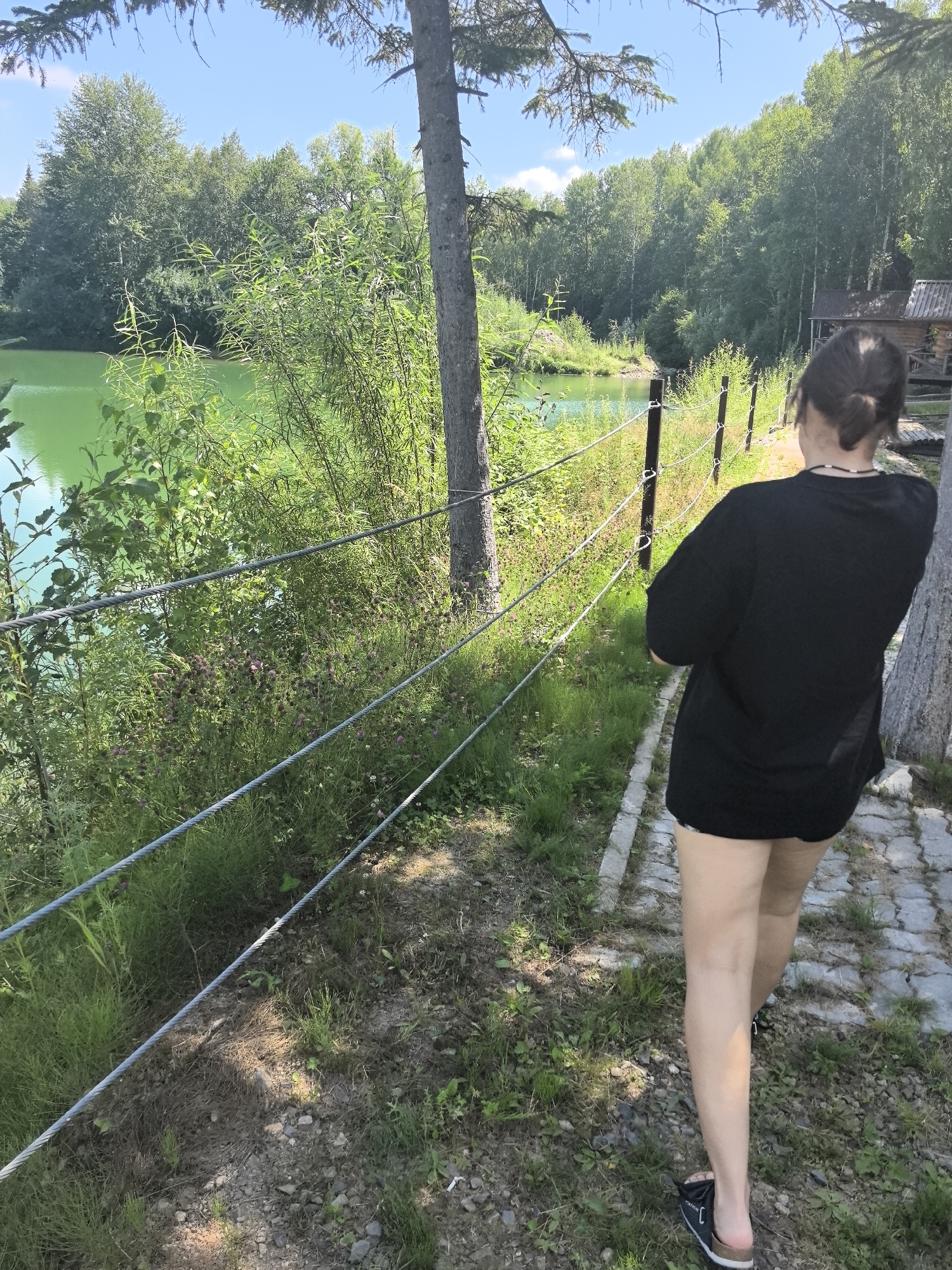 An over-the-shoulder, medium shot shows a young woman walking away from the camera on a cobblestone path. She is wearing a black t-shirt and black shorts, and her hair is pulled back in a bun. The path is bordered on the left by a wire fence and lush green vegetation, and on the right by trees and a rustic wooden cabin. A vibrant green body of water is visible beyond the fence. The scene is set in a natural outdoor environment, likely near a lake or river, with a clear blue sky and scattered clouds overhead. The location is described as Solnechnyi, Russia.