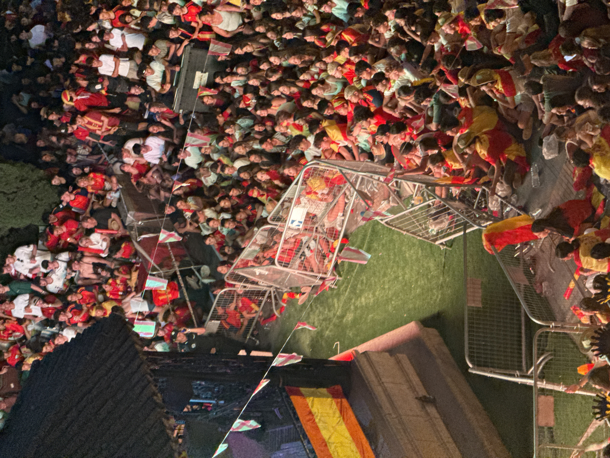 The image shows a large crowd of people, many of whom are wearing red and yellow, gathered in an outdoor area. The crowd appears to be seated on the ground and behind metal barriers. Some individuals are holding flags that are red and yellow, suggesting a possible national or team allegiance. In the background, there is a structure with a dark, angled roof, possibly a stage or building. A Spanish flag is visible on the left side of the image. The ground is covered in artificial turf. The scene suggests an event or gathering, possibly a sporting event, festival, or concert, taking place in Las Rozas de Madrid, Spain. The lighting indicates it is either late evening or night.
