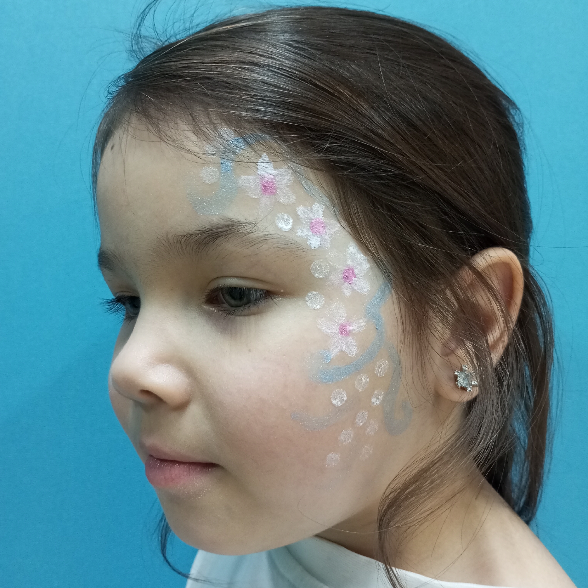 A young girl with dark brown hair is pictured in profile against a solid light blue background. Her face is adorned with a face painting design featuring white flowers with pink centers and blue-grey shimmering accents. She wears a small, shiny earring in her left ear. The image does not contain any details that would place it in Pervomaysky, Russia, or any other specific location. The scene consists solely of the girl and the backdrop.