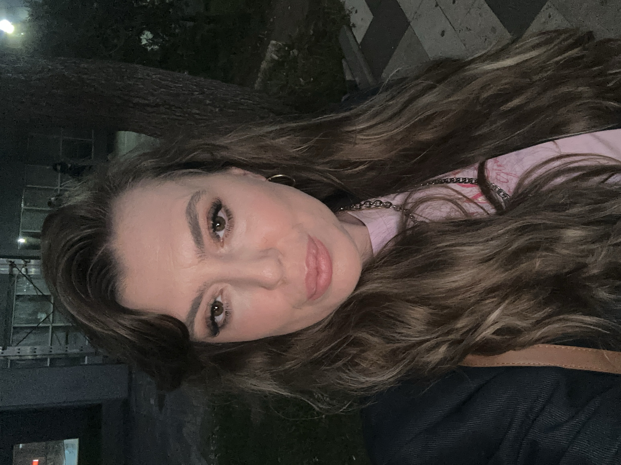 A medium shot depicts a woman with long, wavy brown hair, gold hoop earrings, and a pink t-shirt with a silver chain necklace. She wears dark eye makeup and has light skin. Her expression is neutral, with a slight smile. She is wearing a dark jacket with a brown strap.
The background features a tree trunk on the left side. Behind her head, there is a building façade with visible structural elements and possibly windows. To the right of the building, there is some greenery, and on the right, a tiled pavement is seen. Based on the architectural style and the tiled pavement, the location is likely in an urban area of Batumi, Georgia. The lighting suggests it may be nighttime or a poorly lit area.