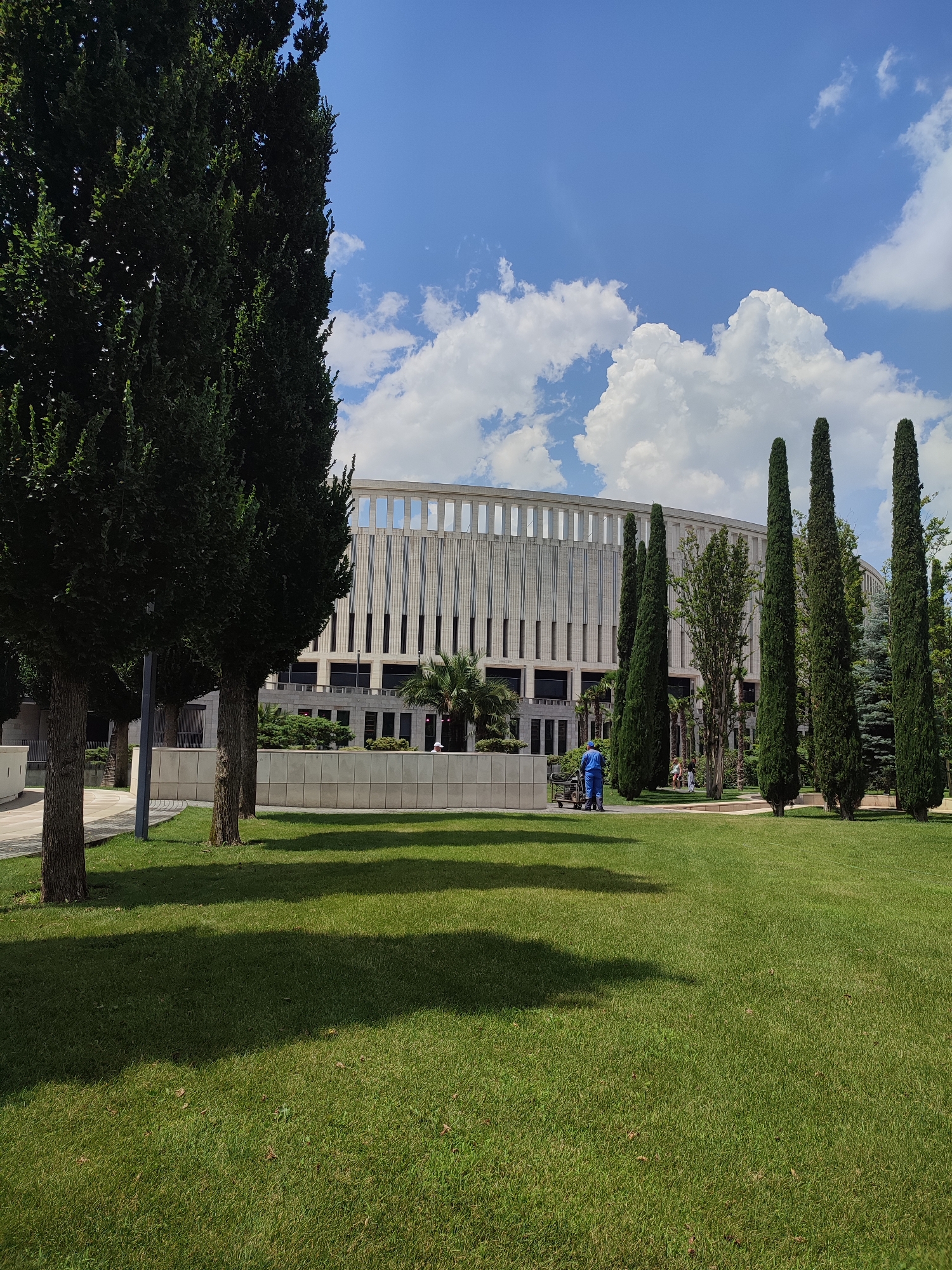 This media file depicts a sunny day in a park-like setting, with a modern building in the background. The foreground is dominated by a well-maintained green lawn, with a few scattered dry leaves. Several tall, dark green cypress trees are planted in a line, creating a natural barrier between the lawn and the building. Other trees, including some deciduous trees with fuller foliage, are also visible.
In the middle ground, a person in a blue uniform is operating a wheeled cart, possibly engaged in gardening or maintenance work. A few other people can be seen further back, sitting or walking in the distance near some trees and bushes.
The building in the background is a large, modern structure with a light-colored facade featuring a repeating pattern of vertical elements and dark windows. Its architecture suggests it could be a public building or administrative complex. The sky above is bright blue with scattered white, puffy clouds, indicative of pleasant weather and daytime. The shadows cast by the trees on the lawn suggest it is mid-day, with the sun relatively high in the sky.
The scene appears to be a public space in Krasnodar, Russia, combining manicured landscaping with contemporary architecture. There is no visible text in the image. The overall mood is calm and orderly, suggesting a well-kept urban environment.