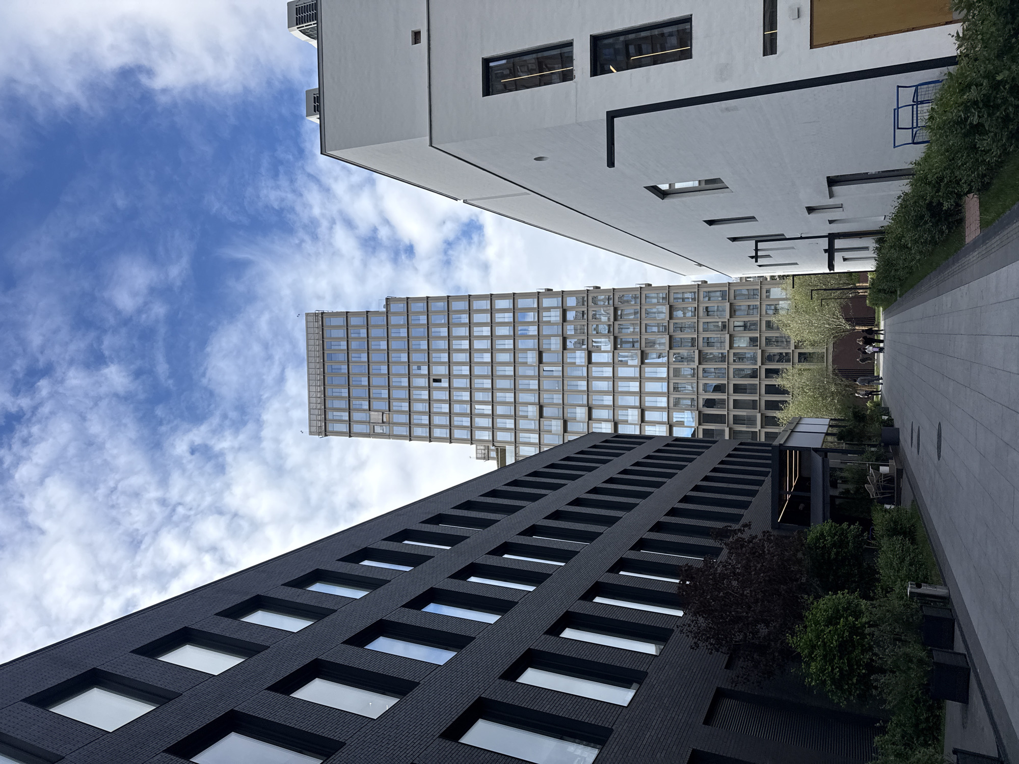 The image shows a street-level view in a city with a concentration of modern high-rise buildings. The sky is light blue with scattered white clouds. Three buildings are prominently visible.
The first building is a dark, possibly black, modern building with a regular pattern of square windows. A second building is tall and narrow, with a grid-like facade of windows and a metal exterior. The third building is white with rectangular windows and some design elements.
Between the buildings is a pedestrian walkway with sparse vegetation and people walking. The image is taken from a low angle.
The location is Moscow, Russia.