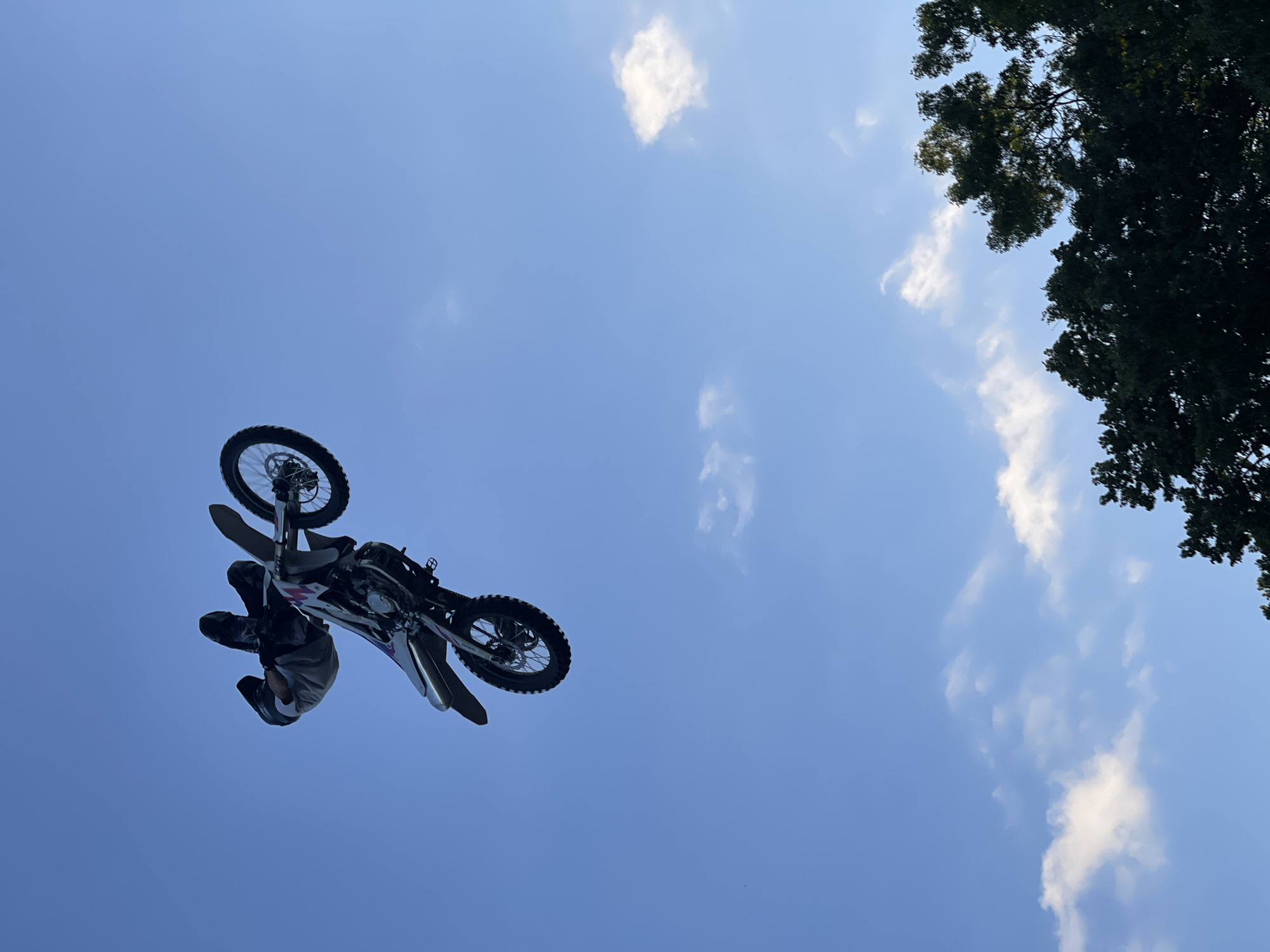A motocross rider performs a stunt mid-air against a clear blue sky with scattered white clouds. The rider is inverted, with their legs bent and body tucked. The dirt bike is also airborne and appears to be performing a flip. The bottom of the frame shows the tops of dark green trees. The image appears to have been taken during daylight. There is no information in the image to indicate a specific location in Strathroy-Caradoc, Canada.