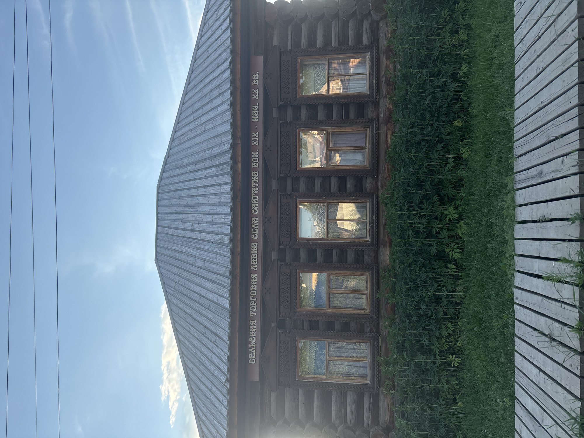 This media file is a photograph of a historic wooden building with several windows and decorative trim. The building appears to be a traditional Russian log house. A sign above the windows reads, in Russian, "СЕЛЬСКИЯ ТОРГОВИЯ ЛИВКИ СЕЛИ САЙГАТКИ КОН. XIX - НИЧ. ХХ ВВ." which translates to "Rural Trade Livka Village Saigatki End of 19th - Beginning of 20th Century." This indicates the building was likely a general store or trading post from that historical period.
The building is surrounded by lush green vegetation, suggesting it is a rural or semi-rural setting. To the right of the building, there is a wooden walkway or boardwalk. The sky in the background is blue with some scattered clouds and visible power lines, implying an outdoor scene during daylight. The sunlight casts reflections on the window panes. The overall mood of the image is one of historical preservation and rustic charm, showcasing a glimpse into the past.