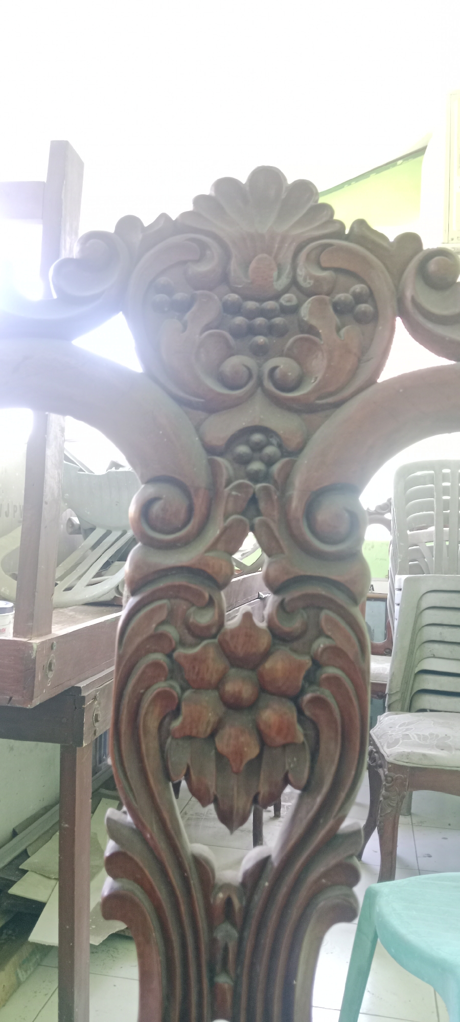The image shows a close-up of an ornate wooden carving, likely part of a chair or furniture. The carving features floral motifs and clusters of what appear to be grapes. The wood is dark brown and shows signs of detailed craftsmanship. In the background, stacked white plastic chairs are visible, suggesting a setting like a hall or an outdoor event space. Some construction materials, possibly plasterboards, are also present in the mid-ground. A portion of a teal plastic stool is visible in the foreground on the right. The lighting suggests an indoor or shaded outdoor environment with bright light from above. The specific location in Tuban, Indonesia, is not visually identifiable from the image itself.