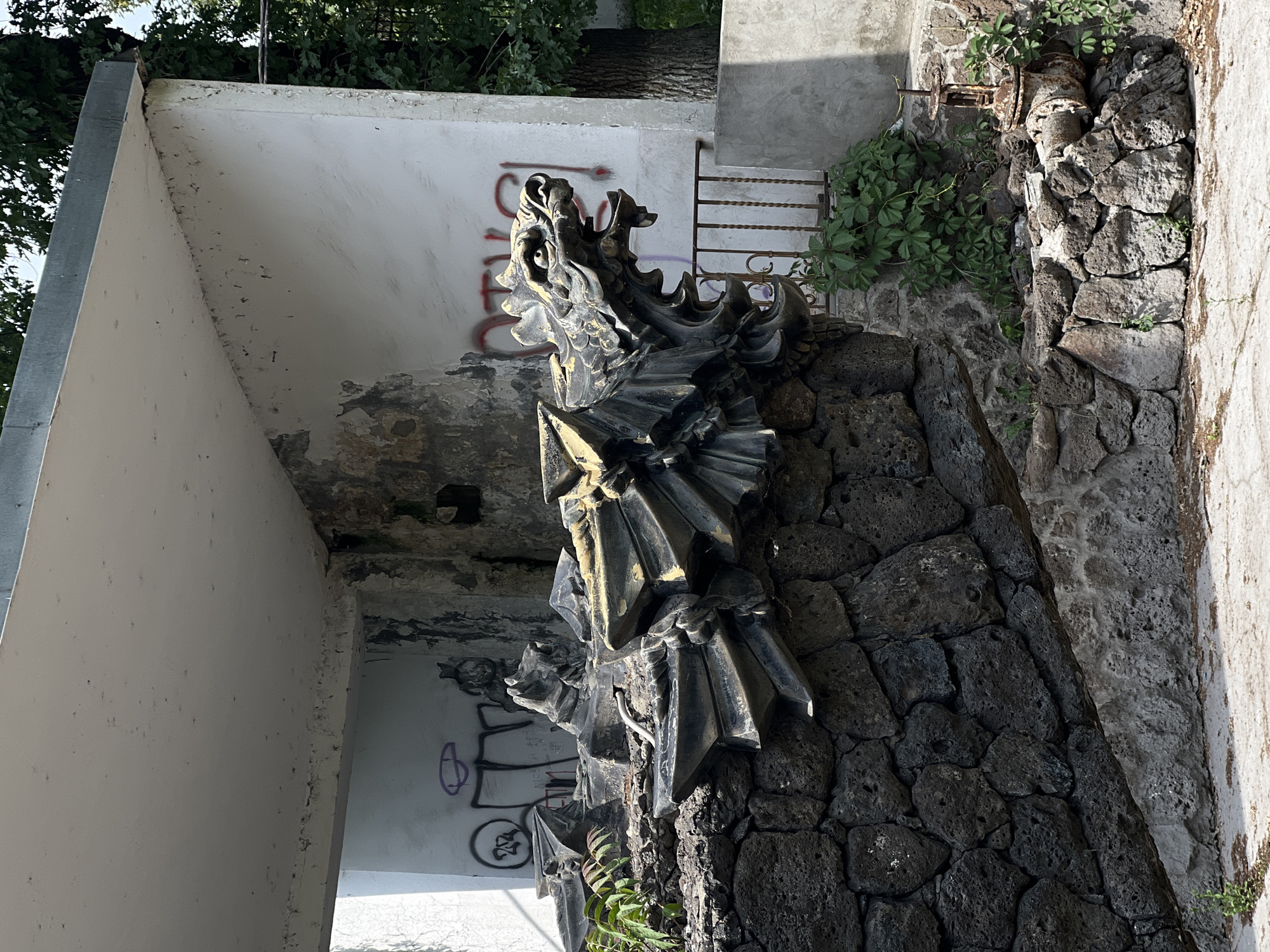 A vertical, medium shot depicts a stone dragon sculpture. The sculpture is affixed to a dark, porous stone wall. Vegetation grows adjacent to the right side of the wall. Graffiti mars a light-colored wall behind the statue, while a metal grate is seen further beyond, also behind the statue. Trees and foliage are visible in the top left of the image. The location is Unknown, Unknown.