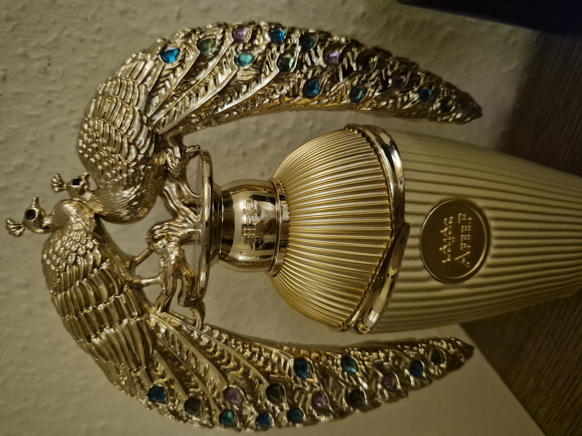 A close-up, eye-level, medium shot captures a golden perfume bottle shaped like a fluted column, topped with two ornate golden peacocks with spread wings. The peacocks' wings are adorned with small, colorful gemstones, including blue, green, and purple. The bottle itself is a bright gold color with vertical ridges. A circular emblem on the lower portion of the bottle displays text that is partially obscured. The background is a textured, light-colored wall, and the object rests on a surface that appears to be wood. The lighting is dim and warm, casting soft shadows. The location is not specified in the image.