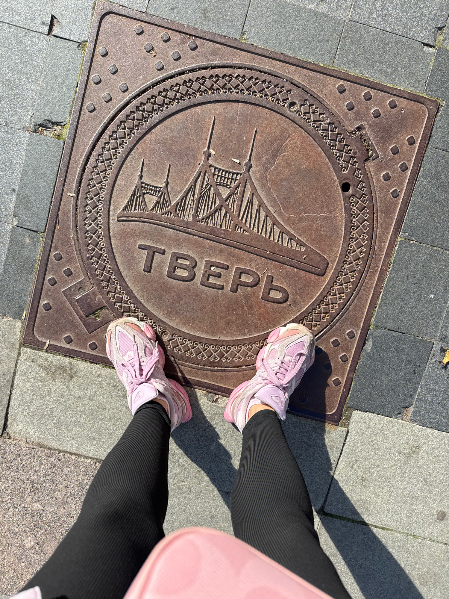The image captures a high-angle, eye-level perspective of a person standing on a sidewalk in Tver, Russia. The person is wearing black ribbed leggings and light pink athletic shoes. Their shadow is cast on the sidewalk to their right.
The central focus of the image is a square manhole cover. The cover is made of dark, weathered metal with raised circular and square patterns around its perimeter. Within the square border, a circular design is prominent. This circular design features an embossed illustration of a bridge with multiple arches and towers. Below the bridge illustration, the word "ТВЕРЬ" (TVER) is inscribed in large, capital letters, indicating the city's name.
The manhole cover is set into a paved sidewalk composed of grey and darker grey rectangular stone slabs. Some of these slabs show cracks and gaps, with small amounts of green moss growing in some of the crevices. The lighting suggests a sunny day, with strong shadows indicating the time of day.