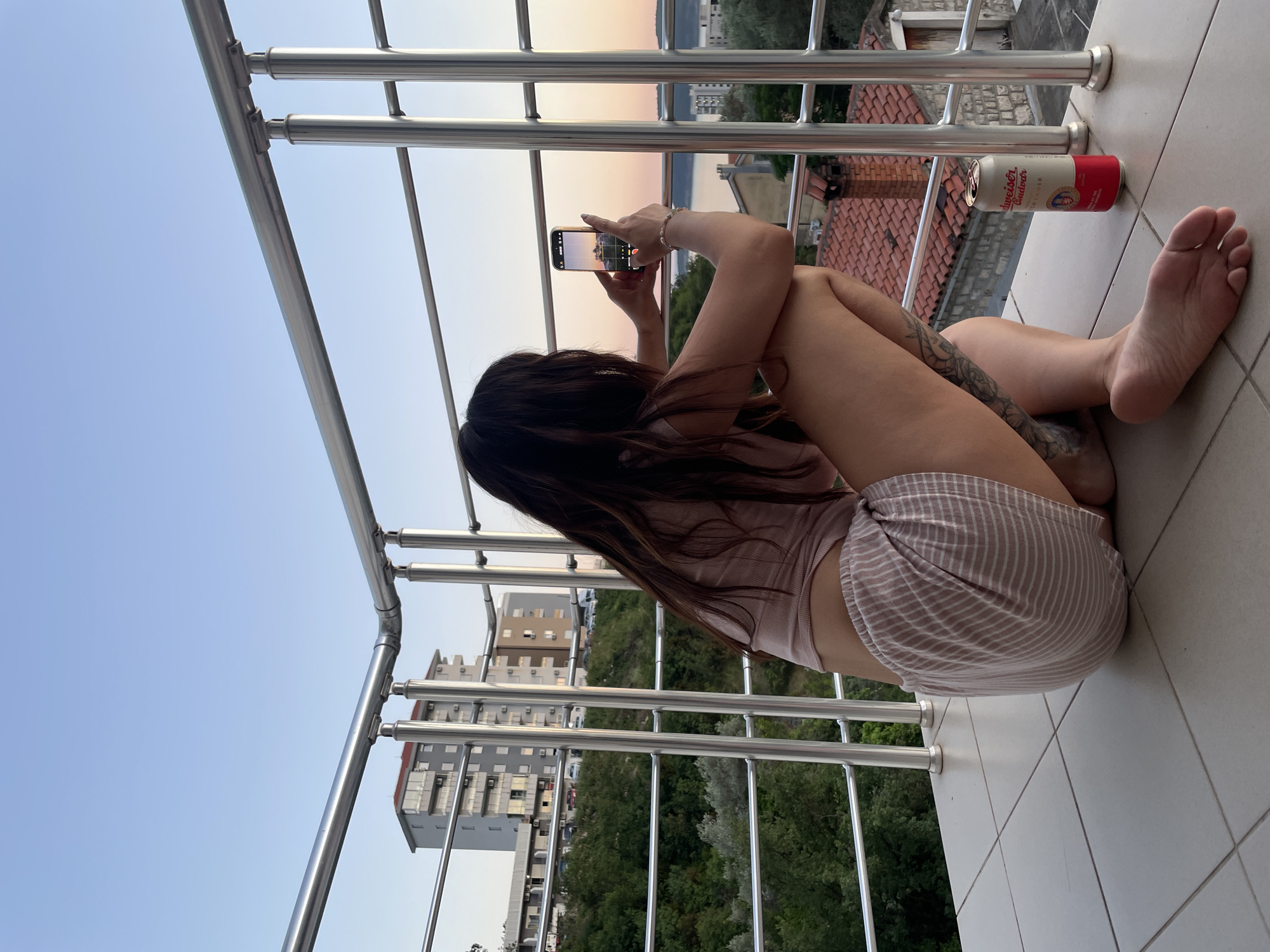 A person with long, dark hair is kneeling on a tiled balcony, holding a smartphone to take a picture of a sunset over the water. They are wearing a light pink cropped top and striped shorts. A can of Budweiser beer is on the balcony floor next to them. In the background, there are buildings, trees, and the sea. The scene appears to be set in Rafailovići, Montenegro, given the context provided. The balcony railing is made of metal. The sky is a gradient of blue and orange, indicating it is dusk.