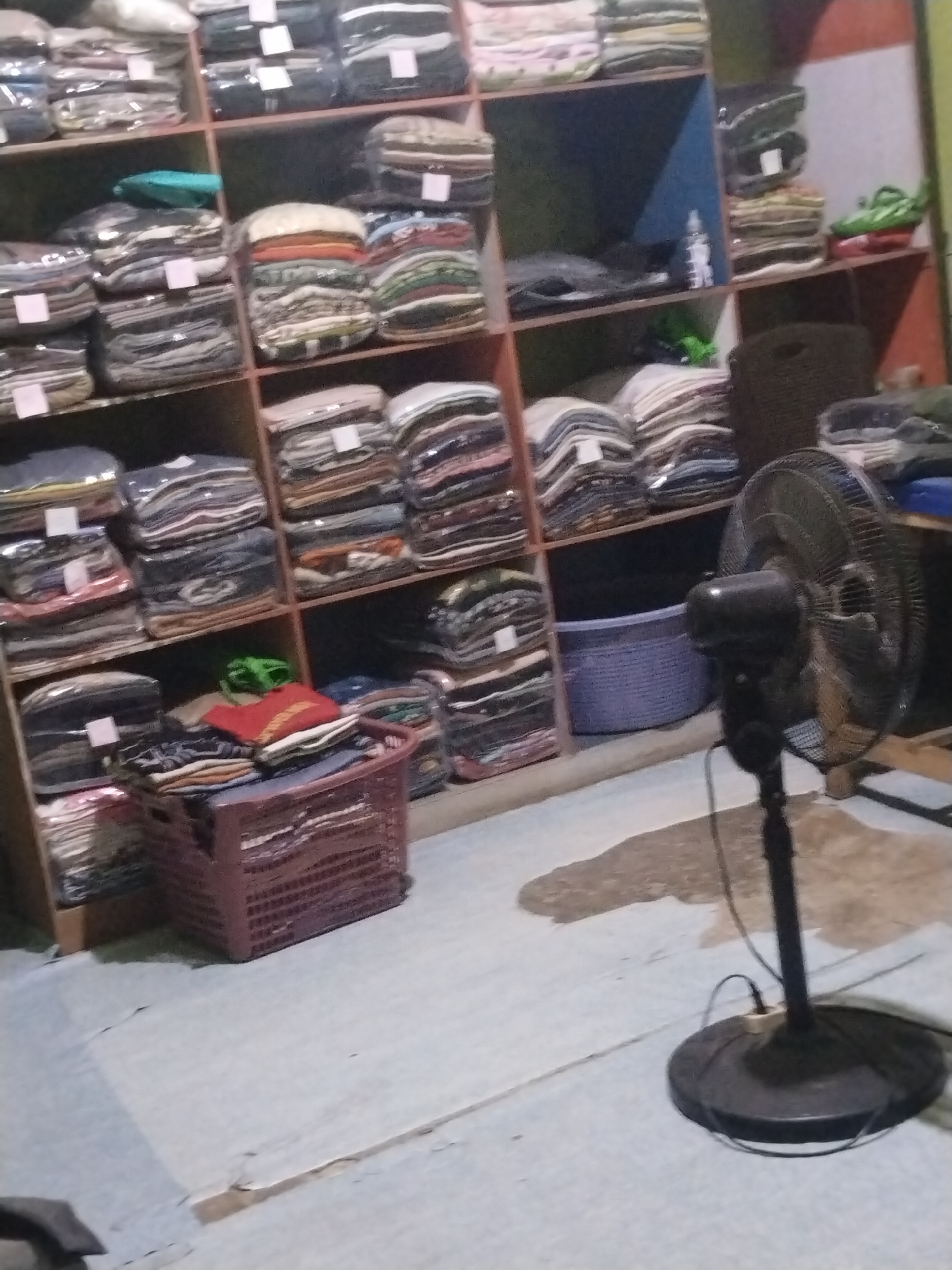 The image depicts a cluttered retail space, likely a small clothing store in an unknown location in Indonesia. Shelves are packed with neatly folded stacks of clothing, many of which appear to be T-shirts or similar garments. The clothing is organized by color and possibly by type. A brown woven basket filled with more folded clothes sits on the floor in the foreground, to the left of a large black standing fan. The fan, which is on, is positioned to the right of the basket and is blowing towards the camera. A purple basket is visible on a shelf behind the fan. The overall impression is one of a working shop with goods for sale, though the lighting is dim, and the image is somewhat blurry.