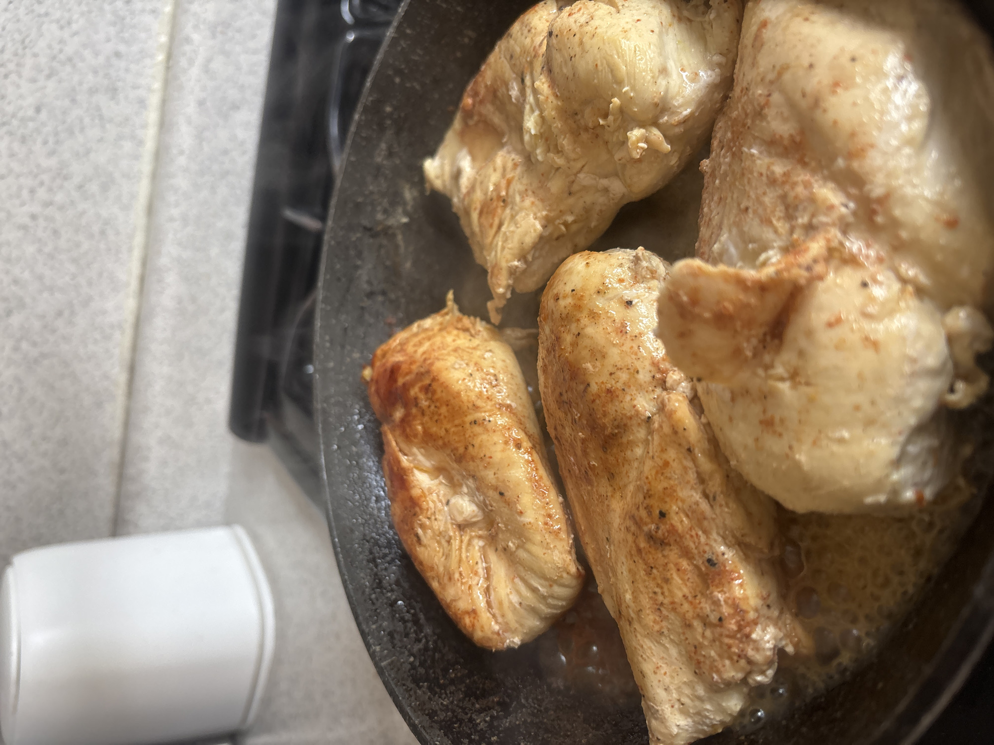 The image captures a close-up, overhead view of chicken breasts cooking in a dark frying pan on a stovetop, likely in a kitchen in Puebla, Mexico. Four substantial pieces of chicken are visible in the pan, actively sizzling in bubbling liquid, suggesting oil or butter. The chicken pieces appear to be skinless and are seasoned with visible reddish-brown and dark speckles, indicating spices. Some parts of the chicken show a golden-brown sear, particularly on the piece towards the bottom left, while others are still lighter in color, suggesting they are in the process of cooking.
The pan itself is dark, possibly cast iron or a non-stick material, and shows some wear. In the background, out of focus, are black grates of what appears to be a gas stove. To the left of the pan, a light grey, textured countertop or kitchen surface is visible. In the bottom left corner, a white, cylindrical object with rounded edges, resembling a spice shaker or a small container, lies on its side, also slightly out of focus. The lighting appears to be indoor and artificial, illuminating the food and immediate surroundings. There is no visible text in the image.