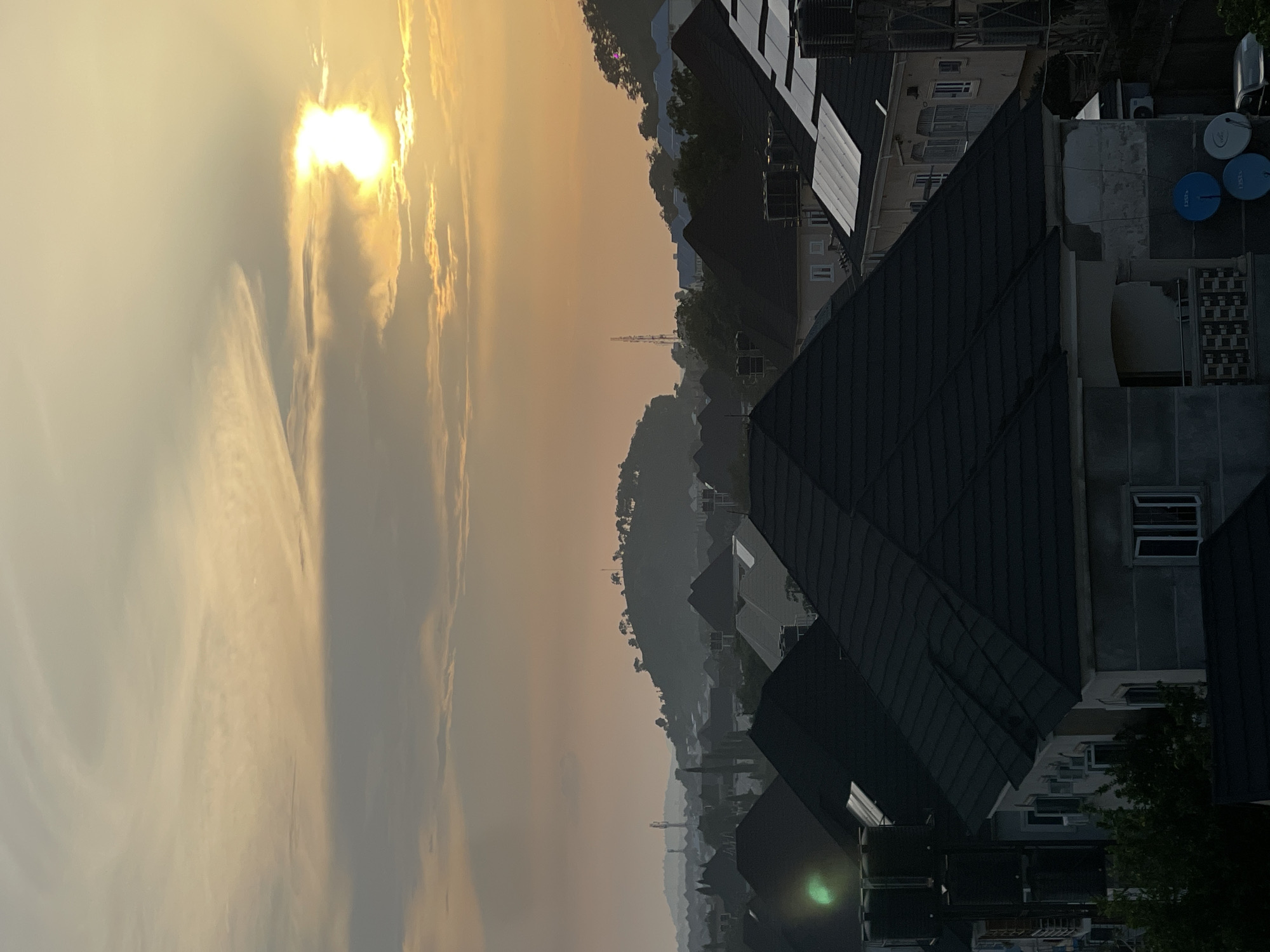 This is an aerial photograph of the city of Abuja, Nigeria, taken at either sunrise or sunset, with the sun prominently visible in the upper right quadrant of the frame. The sky is filled with a soft, warm glow, with streaks of clouds stretching across the frame, illuminated by the setting or rising sun. Below the sky, a hilly landscape dominates the background, dotted with trees and what appear to be communication towers. In the foreground, a densely packed residential area is visible, characterized by numerous houses with dark-colored roofs. Some of these roofs are adorned with satellite dishes and solar panels. The buildings are of varying sizes and architectural styles, suggesting a mix of modern and older constructions. The overall impression is of a sprawling urban environment nestled within a natural, hilly landscape.