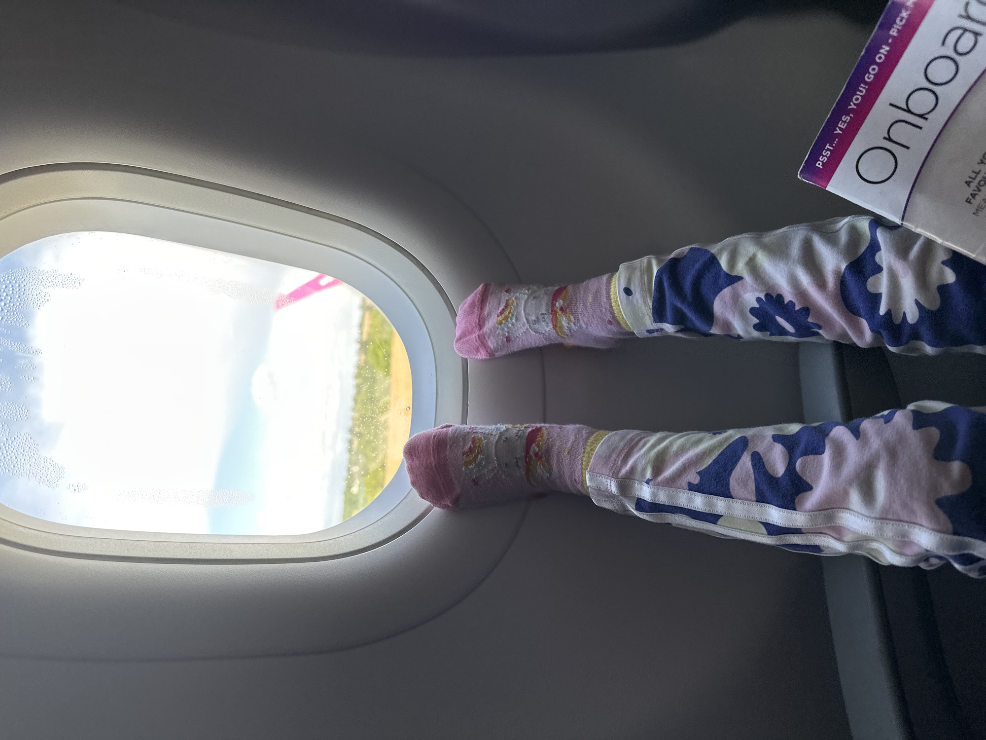 This image shows a view inside an airplane with a child's legs and feet propped up against the window. The child is wearing patterned pajamas and pink socks with cartoon graphics. The airplane window shows a landscape outside with a blue sky, white clouds, and a glimpse of land. Beside the legs, there's an "Onboard" magazine. The window has some condensation on it.