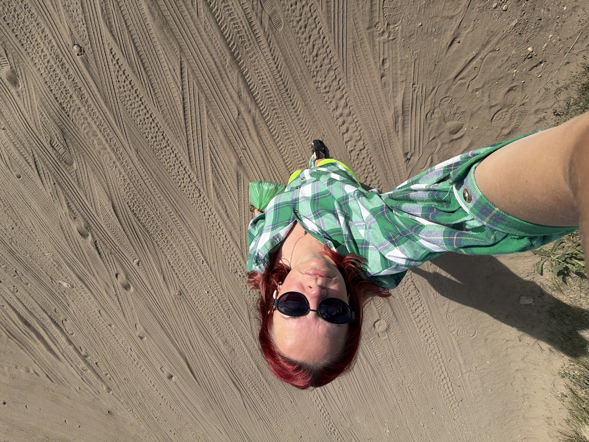 A woman with red hair, wearing sunglasses and a green plaid shirt, takes a selfie while standing on a sandy surface covered with tire tracks. The location is likely an outdoor area in or near Chaikovsky, Russia, given the request for context. The woman is holding a green plastic bag in one hand.