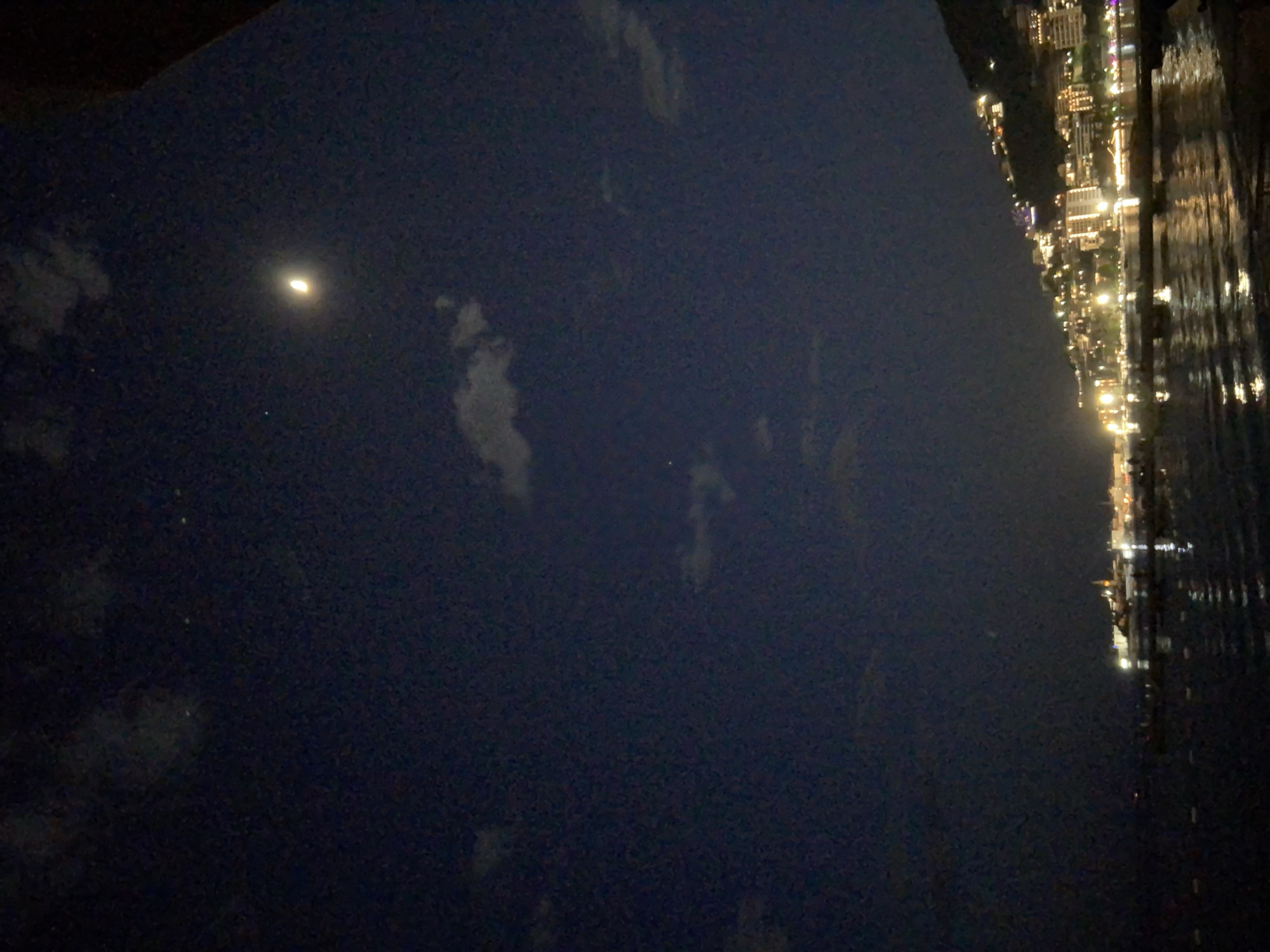 A nighttime image shows a body of water and the edge of a cityscape. The body of water takes up the left half of the image, with a crescent moon and some faint clouds visible in the sky. The city lights illuminate the right side of the image, and their reflections are visible in the water. The city appears to be located along a coastline.
