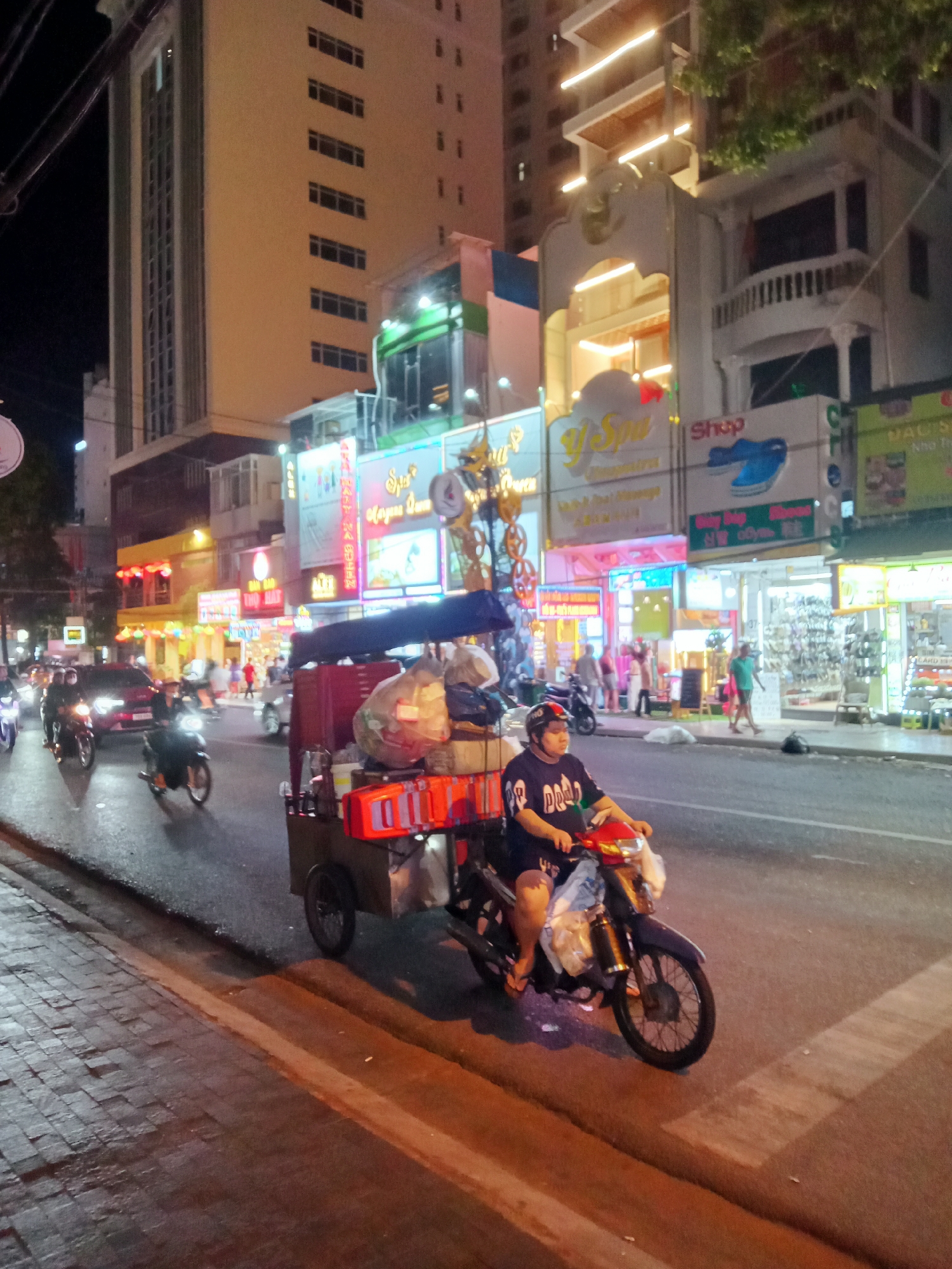 This vibrant night scene captures a bustling street in Nha Trang, Vietnam, illuminated by numerous artificial lights from shops and street infrastructure. Tall buildings line both sides of the road, their windows glowing, while commercial establishments on the ground floor display an array of colorful neon signs and brightly lit interiors.
The primary subject is a person, likely male, wearing a helmet and a dark t-shirt with partially visible text like "DOD," driving a unique motor-tricycle. This vehicle consists of a red/maroon scooter attached to a large, boxy cargo cart loaded with plastic bags, possibly containing goods, and several orange crates. The driver appears to be navigating the busy street.
Other vehicles, including several motorbikes and at least one red car, are also in motion on the road, creating a sense of active urban traffic. People are visible further down the street, walking on the sidewalks and near the brightly lit shopfronts, suggesting commercial and social activity.
Notable details include the bright, almost overwhelming, artificial lighting that defines the scene, creating reflections on the wet-looking asphalt. Overhead power lines are visible in the upper left. Shop signs prominently feature "Y Spa" in yellow script, and another sign reads "Shop" with a blue shoe icon, accompanied by Korean characters (신발, meaning shoes). The overall impression is one of a lively, working city street at night.