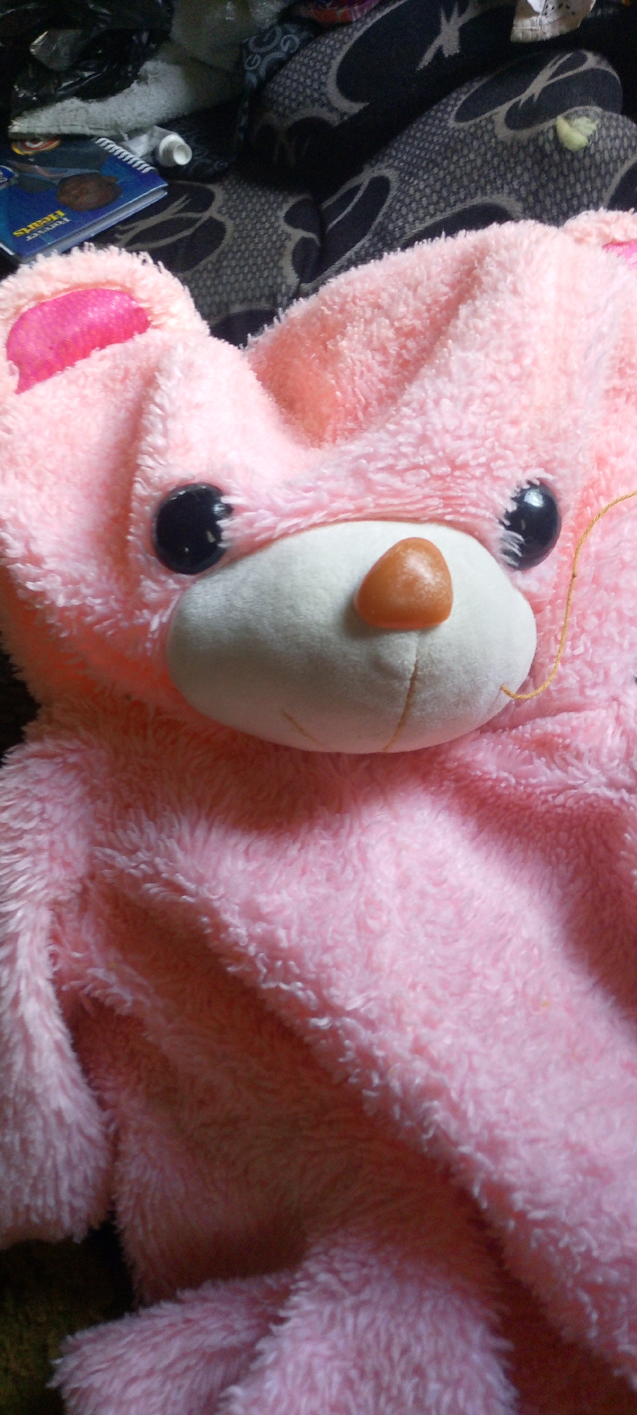 The image primarily features a large, fluffy, pink plush toy, likely a teddy bear or similar animal. The plush toy's head and upper body occupy most of the frame, showcasing its face with prominent glossy black eyes, a soft white snout, and a small, reddish-brown, triangular nose. One pink inner ear is partially visible on the left side of its head. The entire visible surface of the plush toy is covered in soft, shaggy pink fur, emphasizing its plush texture. A thin, yellowish string or thread extends from the right side of the plush toy's snout.
In the blurred background, particularly in the upper left corner, several objects are visible amidst darker, patterned fabrics, which could be blankets, clothing, or furniture upholstery. These objects include a small blue spiral-bound notebook, partially displaying the text "Forever Hearts," a small dark, irregularly shaped item (possibly metallic), and a white tube.
The setting appears to be indoors, possibly a domestic environment such as a bedroom or living area, given the nature of the subjects. There is no specific activity or event taking place; the plush toy is stationary. There are no clear indicators of the time of day or weather. The image does not contain any specific visual cues to confirm the stated location of Adankolo, Nigeria, as the scene is generic.