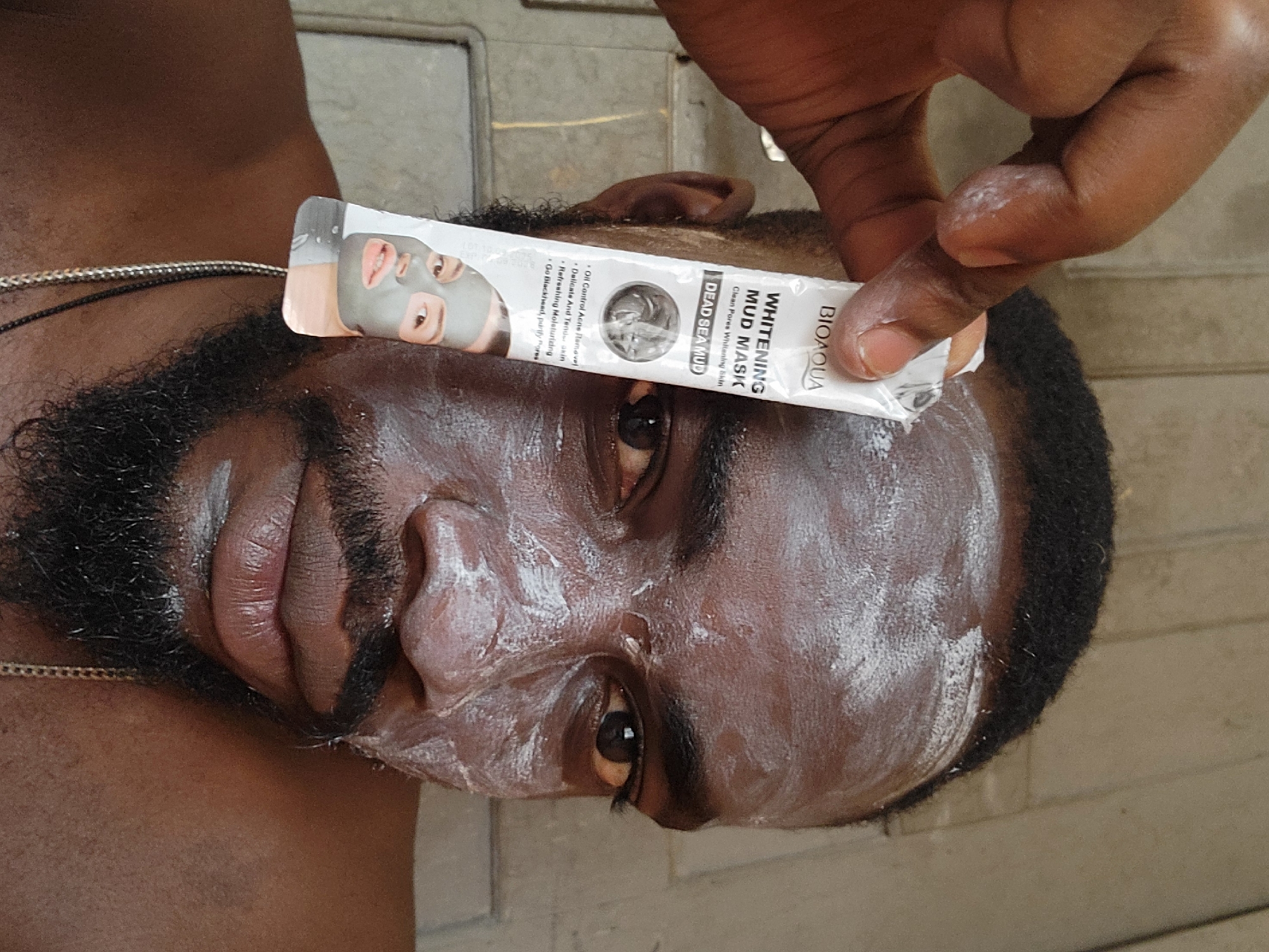 A close-up, rotated image shows a Black man with a mud mask applied to his face, specifically around his nose and cheeks. He has a dark beard and short, dark hair. A hand holds a sachet of Bioaqua Whitening Mud Mask just above his eyes, partially obscuring them. The sachet features an image of a person wearing a grey mask, and text that is partially readable: "WHITENING MUD MASK, Clean Pores Whitening Skin, DEAD SEA MUD, Oil Control Acne Remove, Delicate And Tend skin, Refreshing Moisturizing, Go Blackhead, purify Fores Pro." The background features a tiled surface.
There is no information available to suggest a specific location within the city of Jos, Nigeria.