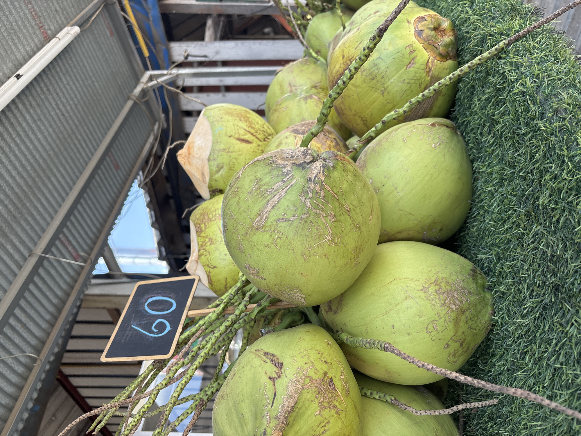 A bunch of green coconuts, seemingly for sale, are stacked together with visible stems. A small, black chalkboard with the price "60" written in blue is stuck into the bunch. The coconuts rest on a patch of artificial green turf. The background reveals parts of a metal structure, possibly a market stall or shop roof, with sunlight glinting off metal elements. The location is Choeng Thale, Thailand.