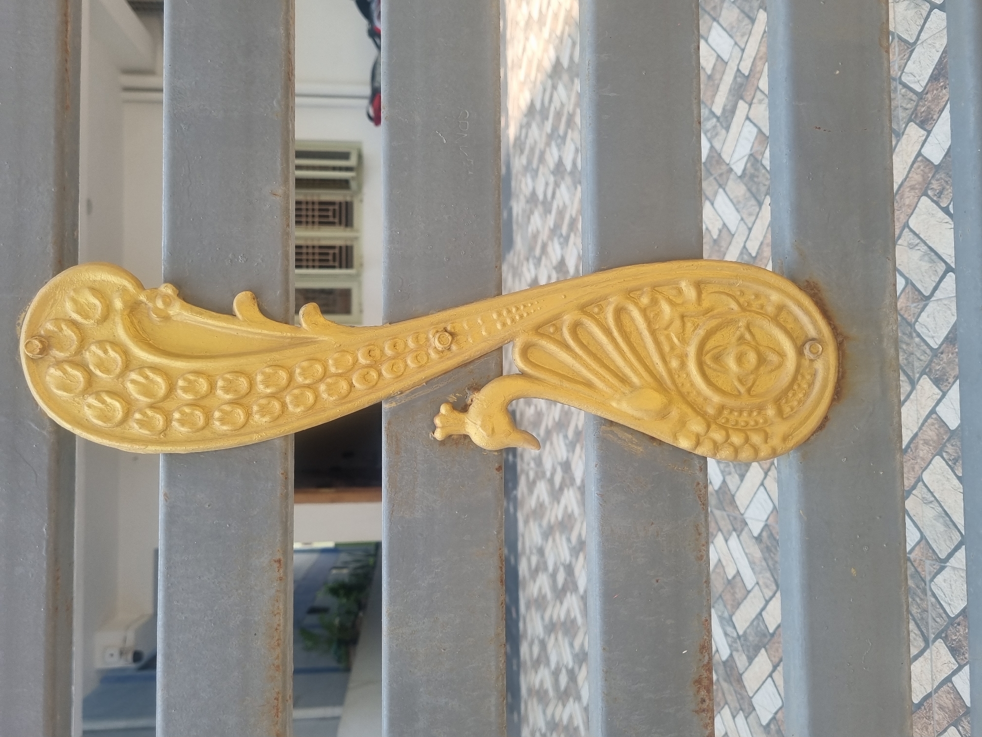 An eye-level, outdoor, close-up shot shows a gold-colored ornamental element attached to a grey metal gate in Pedda Kammavaripalli, India. The gate is composed of vertical bars. The background shows sections of flooring or paving stones and partial views of buildings. The ornamental element appears to be shaped like an elongated paisley or a stylized peacock. The element has circular details on one end, a small bird figure in the middle, and floral-like patterns on the other end. The element is attached to the gate with fasteners. There is slight surface rust visible on the metal gate bars.