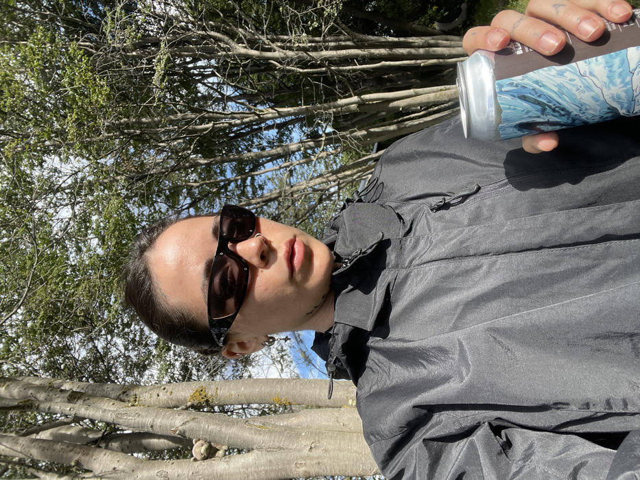 A person wearing sunglasses and a black jacket is holding a can, possibly a beverage, in their right hand. The person has dark hair pulled back and a nose ring. Tree branches and sky are visible in the background, suggesting an outdoor setting. The image is tilted, with the person's face angled towards the camera. The overall lighting appears to be bright, possibly from natural sunlight.