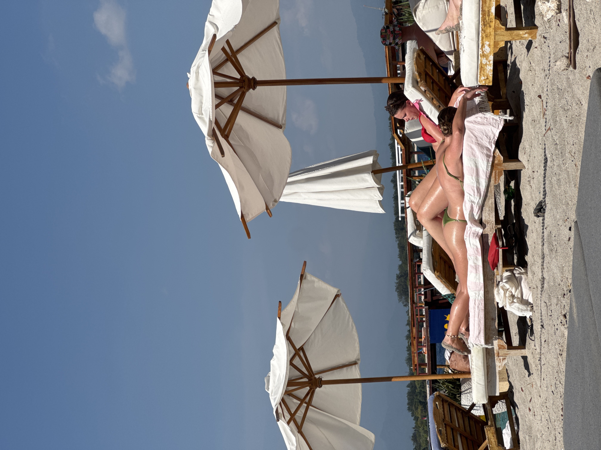 Two women are sunbathing on lounge chairs on a sandy beach under white umbrellas. The woman in the foreground is lying on her stomach wearing a green bikini, her body is oiled and glistening in the sun. The woman behind her is sitting up and wearing a red top. In the background, there are trees, mountains, and a clear blue sky. The scene is likely in Indonesia given the presence of palm trees in the distance.