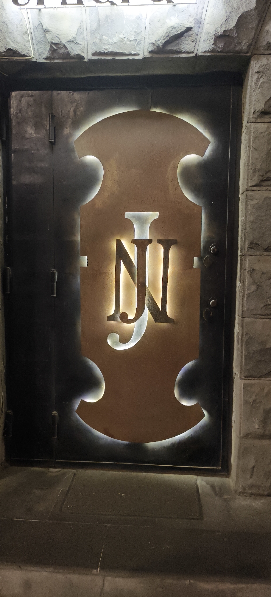 A dark, heavy metal door, likely an entrance to a building, is featured in a close-up, eye-level shot. The door is set into a wall constructed from large, light-grey, rough-hewn stone blocks. The dominant element on the door is a prominent, illuminated sign. This sign has a warm, bronze-toned back panel, shaped distinctively like the body of a string instrument, such as a violin or cello.
Within this illuminated panel, a stylized, dark monogram of the letters "NJ" is centrally placed. The letters themselves are also back-lit, creating a soft, bright glow around their edges and highlighting their ornate design. The illumination suggests it is nighttime or late evening, as the surroundings are dim.
Above the door, slightly out of focus, is another sign with text that appears to read "OPERA" in a decorative, gothic-style font. The ground directly in front of the door consists of dark, rectangular paving stones. The overall scene conveys a sense of a sophisticated or exclusive establishment, possibly a bar or restaurant, in an urban setting like Yerevan, Armenia. There are no people visible, and no specific activity is taking place.