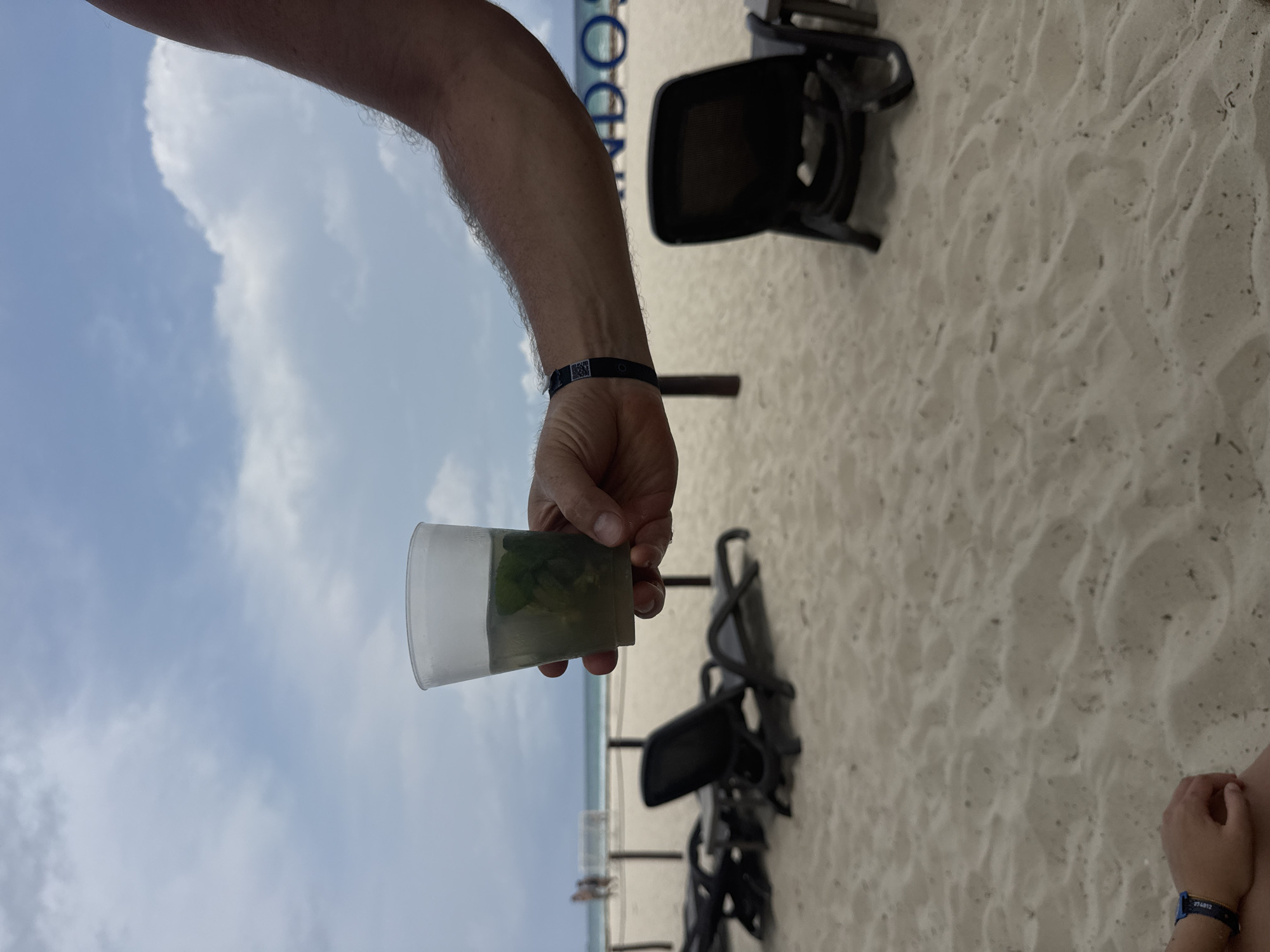 A vertical shot shows a person's arm holding a cup of a green liquid against a blue sky with clouds in Playa del Carmen, Mexico. The background is a beach with lounge chairs in rows. A small child's hand is visible on the lower right.
