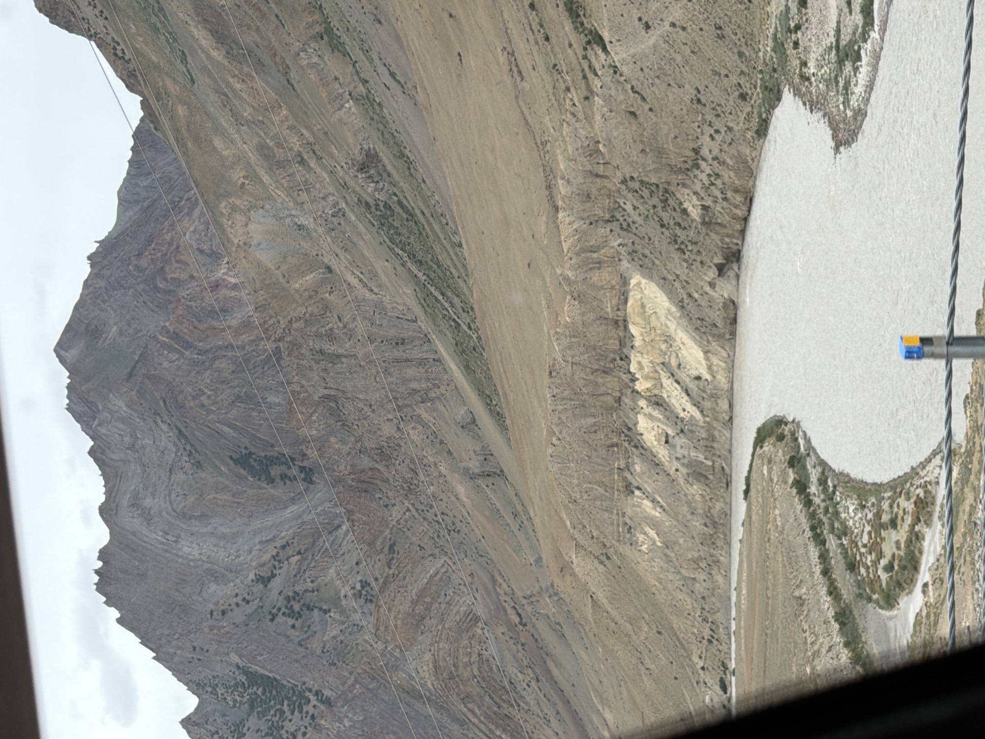 A wide, muddy river flows through a valley with steep, rocky mountains on either side. The mountains are layered with different shades of brown and gray rock, showing signs of erosion and geological folding. Sparse vegetation, appearing as small green shrubs and patches of dry grass, clings to the slopes. In the foreground, a portion of a dark frame, possibly from a vehicle window, obscures the lower left corner of the image. Several thin, parallel wires stretch across the lower part of the frame, with a small blue and yellow marker visible on one of them. The sky is overcast and a pale gray. The scene depicts a natural landscape with no visible human activity or structures, suggesting a remote, mountainous location such as the city of Pidmo, India, known for its dramatic terrain and river valleys.