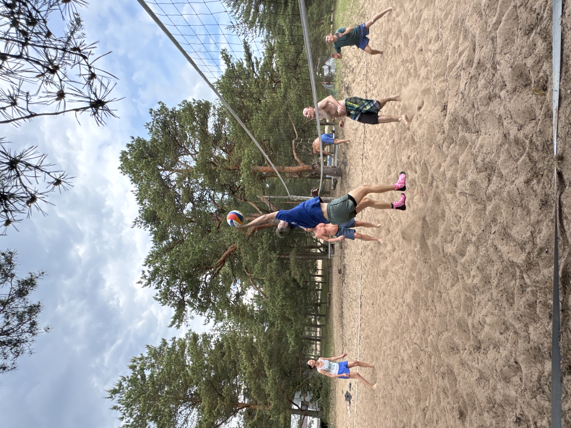 This image depicts an active beach volleyball game in Zelenaya Roshcha, Russia, under an overcast sky. Six individuals are engaged in the sport on a sandy court, which is bordered by dense pine forest.
In the foreground, a male player, wearing a blue t-shirt, green shorts, and prominent pink and black patterned socks, is captured mid-jump, positioned to spike a multi-colored volleyball with his right hand over the net. To his left, a female player, dressed in a white and blue patterned tank top and blue shorts, stands observing the play, facing the central player. To his right, a shirtless male player in plaid swim shorts stands with arms slightly outstretched, also observing the action. Further to the right, a fourth male player in a dark green t-shirt and blue shorts is crouched, ready. In the background, behind the central player, another shirtless male player in blue shorts is partially visible. A sixth male player, shirtless and in blue shorts, is visible further back on the left side of the court among the trees.
The volleyball net stretches across the middle ground of the image. The ground is composed of light brown sand, showing textured ripples and footprints from the players. A white court line is visible on the left side of the sand. Tall, dark green pine trees with rugged trunks form a dense backdrop, encompassing the playing area. In the far left background, a portion of a white building or structure is visible through the trees. The sky above is largely cloudy, with patches of lighter grey. Dark pine branches are partially visible in the very top corners of the frame. A pair of dark shoes and some folded clothes rest on the sand near the left court line.