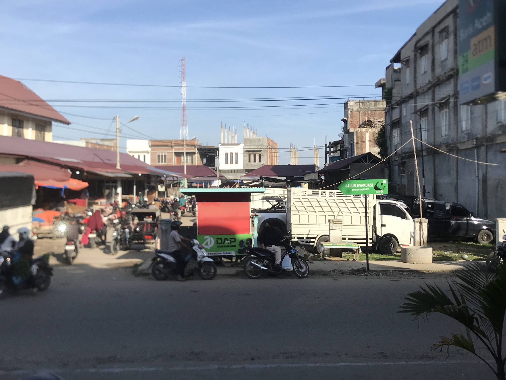 The image depicts a street scene in Lhokseumawe, Indonesia. On the left, there is a covered market area with numerous people and motorcycles, suggesting a busy trading day. Further down the street, construction appears to be in progress on some buildings.
To the right of the street, a small white truck is parked near a sign with green writing. Next to the truck, there is a dark-colored pickup truck parked on a patch of grass.
In the background, there is a red and white communications tower. The sky is blue and relatively clear, indicating fair weather. The overall scene suggests a typical day in a busy, developing urban area.