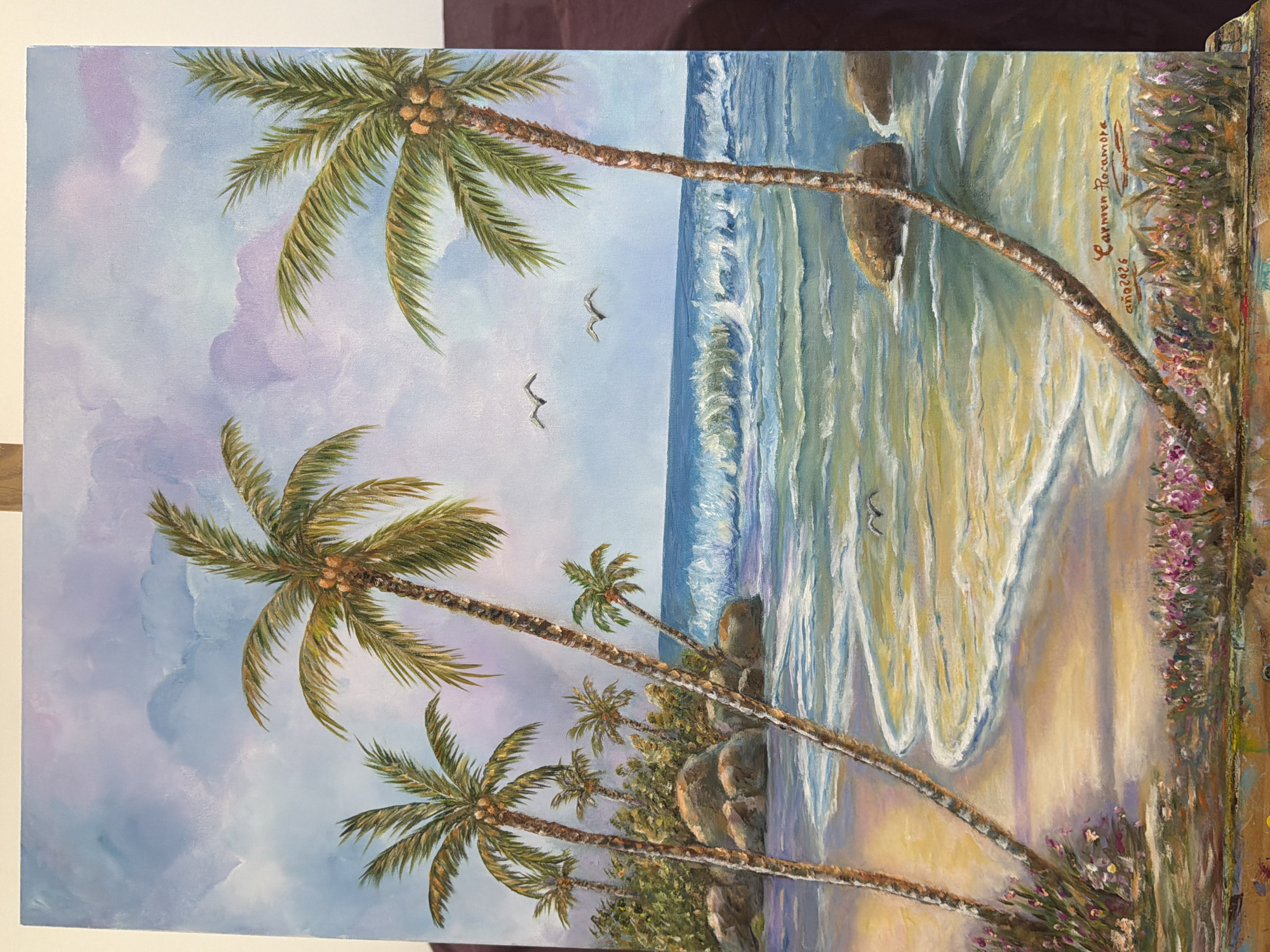 This is an oil painting of a tropical beach scene. In the foreground, there is a sandy beach with clusters of pink and purple flowers. The ocean is depicted with vibrant blue and green hues, showing gentle waves rolling onto the shore. Several palm trees are scattered across the scene, with their fronds reaching towards a sky filled with soft, wispy clouds in shades of blue, purple, and pink. Two birds are shown flying in the distance over the water. The painting is signed "Carmen Rocamora" and dated "año 2026". The painting does not depict any specific location in the city of Elche/Elx, Spain.