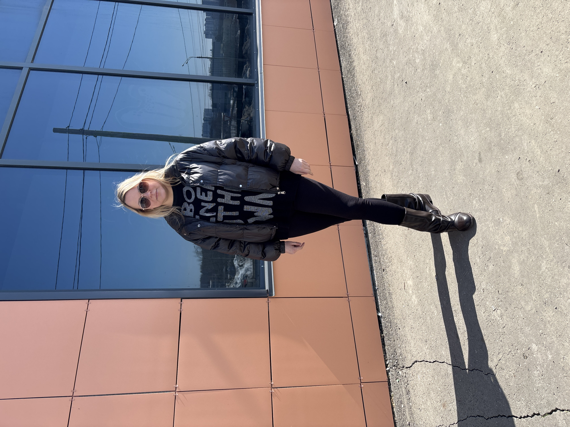An adult female stands on cracked concrete pavement in Izhevsk, Russia, centered before a building facade. The building's left portion features reddish-brown square cladding panels, while the right portion is a reflective glass curtain wall. Visible reflections in the glass include a clear blue sky, a utility pole with multiple overhead wires, distant multi-story buildings, a street light, and remnants of snow on the ground. The subject, a woman with shoulder-length blonde hair and wearing round sunglasses, is dressed in a black puffer jacket, a black long-sleeved top with partial light-colored text ("BO NE TH WA"), black leggings, and dark, mid-calf boots. She stands facing the observer directly, with her right leg crossed over her left ankle. Strong sunlight from the left casts her elongated shadow onto the pavement to her right.