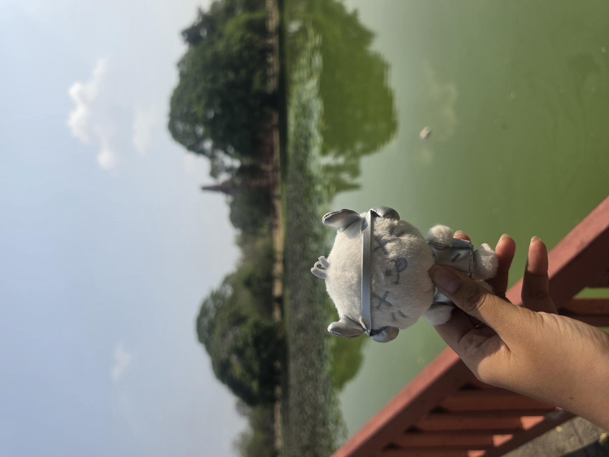 A hand holds a small, plush, grey and white stuffed animal with the letters "X" and "P" embroidered on its body. The background features a green body of water and trees, suggesting a park or natural setting. The scene is likely located in Phra Nakhon Si Ayutthaya City Municipality, Thailand, as indicated by the presence of a partially visible stupa or temple structure among the trees. The sky is light blue with a few clouds, indicating a bright day. The hand is positioned near a red railing. The photo appears to be taken at an angle, potentially to include the water and trees in the background.