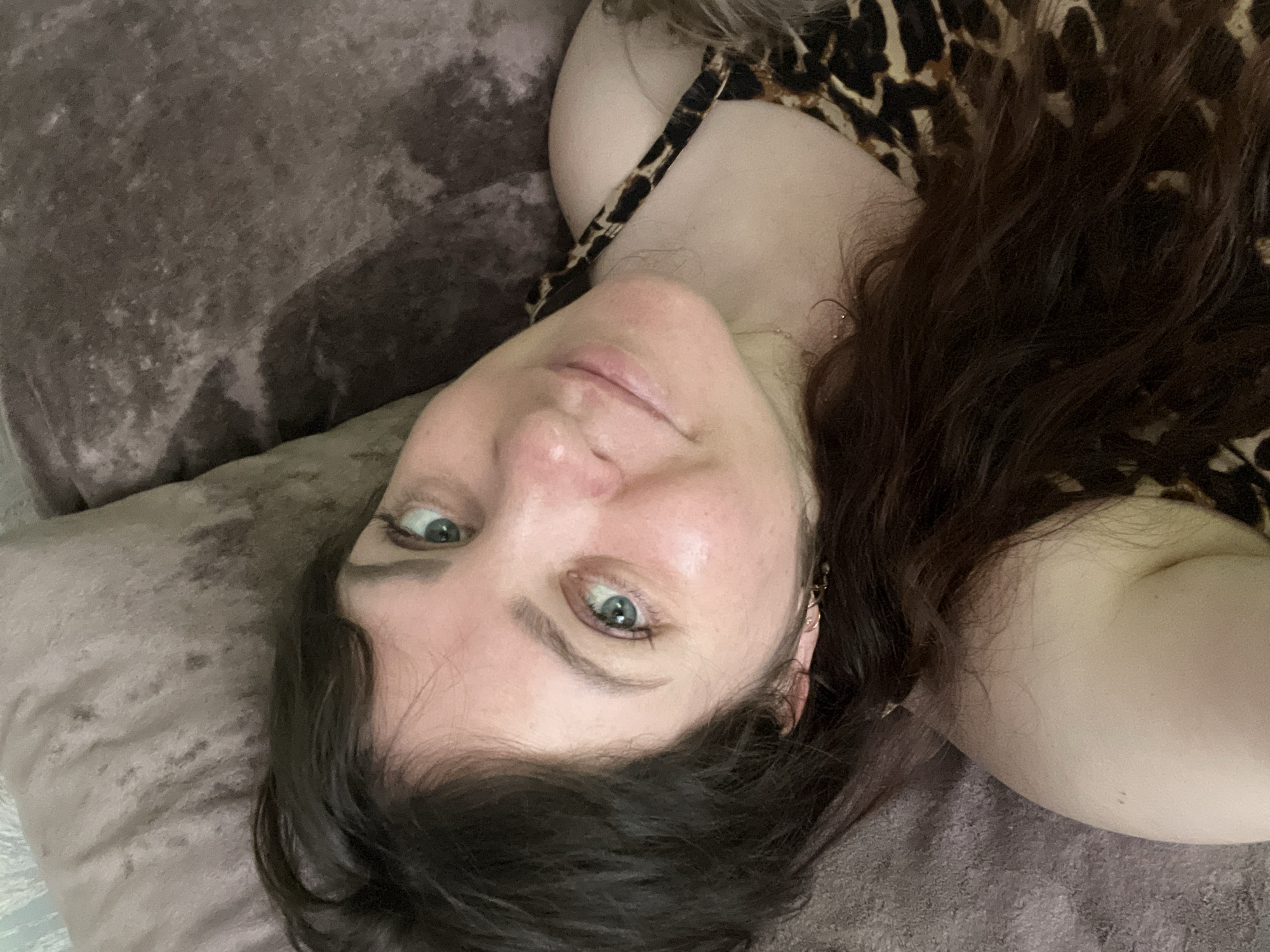 The image is a close-up, horizontal selfie of a woman lying on a textured, brown surface, possibly a couch. She has dark brown hair that is spread out around her face and shoulders. Her head is tilted, and she is looking directly at the camera with light blue eyes. She is wearing a leopard-print top. The lighting appears to be natural, casting soft shadows and highlighting her facial features. The background is out of focus and shows the texture of the surface she is lying on. The setting is indoors, and the image appears to be taken from a high-angle perspective.