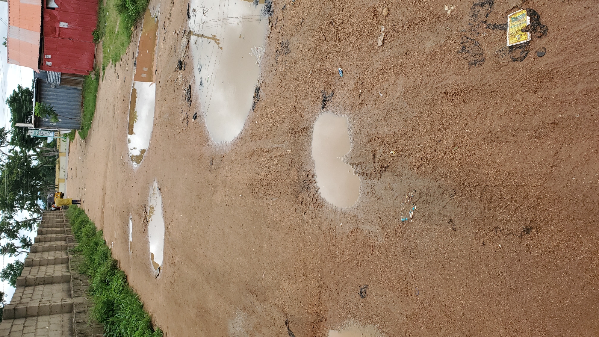The image depicts a dirt road in Jalingo, Nigeria, characterized by numerous muddy puddles and tire tracks. The road surface is predominantly sandy brown, with evidence of recent rainfall. On the left side of the road, a concrete block wall runs along its length, bordered by overgrown green vegetation. Further down the road, a person wearing a yellow garment is walking away from the camera. On the right side of the road, there are corrugated metal structures, including a red building with an orange roof. The background shows a sky with scattered clouds, trees, and some distant structures. The scene suggests a somewhat neglected urban environment.
