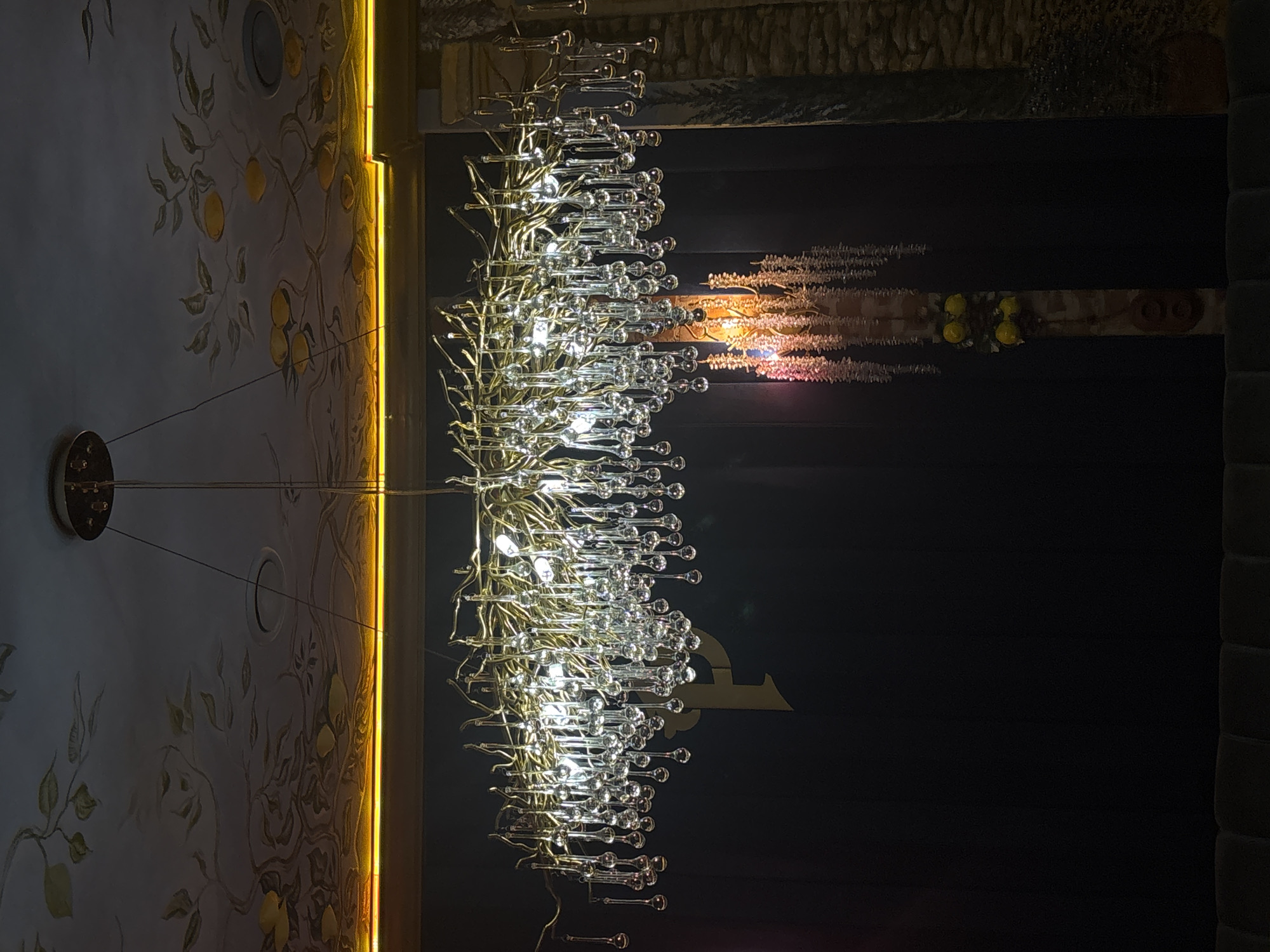 The image depicts a grand chandelier with a complex design, featuring numerous metallic branches and clear glass teardrop-shaped elements. The chandelier is illuminated, casting a soft glow. Above the chandelier, the ceiling is decorated with an artistic mural of branches and fruit, likely lemons, in muted tones. A strip of warm yellow light runs along the top edge of the wall, providing accent lighting. The background is a dark, solid wall, with a subtle hint of a large, golden decorative element, possibly a stylized letter or emblem, behind the chandelier. To the right, a textured stone wall or pillar is visible, and further to the right, a dark, leafy plant or shrub. In the lower right quadrant, a decorative vertical element with hanging foliage and yellow floral accents is visible, illuminated by a pinkish-purple light. The image appears to be taken from a slightly low angle, looking up at the chandelier and ceiling. There are no people visible in the scene. The setting suggests an interior space, possibly a restaurant, hotel lobby, or a venue for events. While the provided text mentions Minsk, Belarus, this specific location is not visually identifiable from the image itself. The scene conveys an atmosphere of opulence and artistic design.