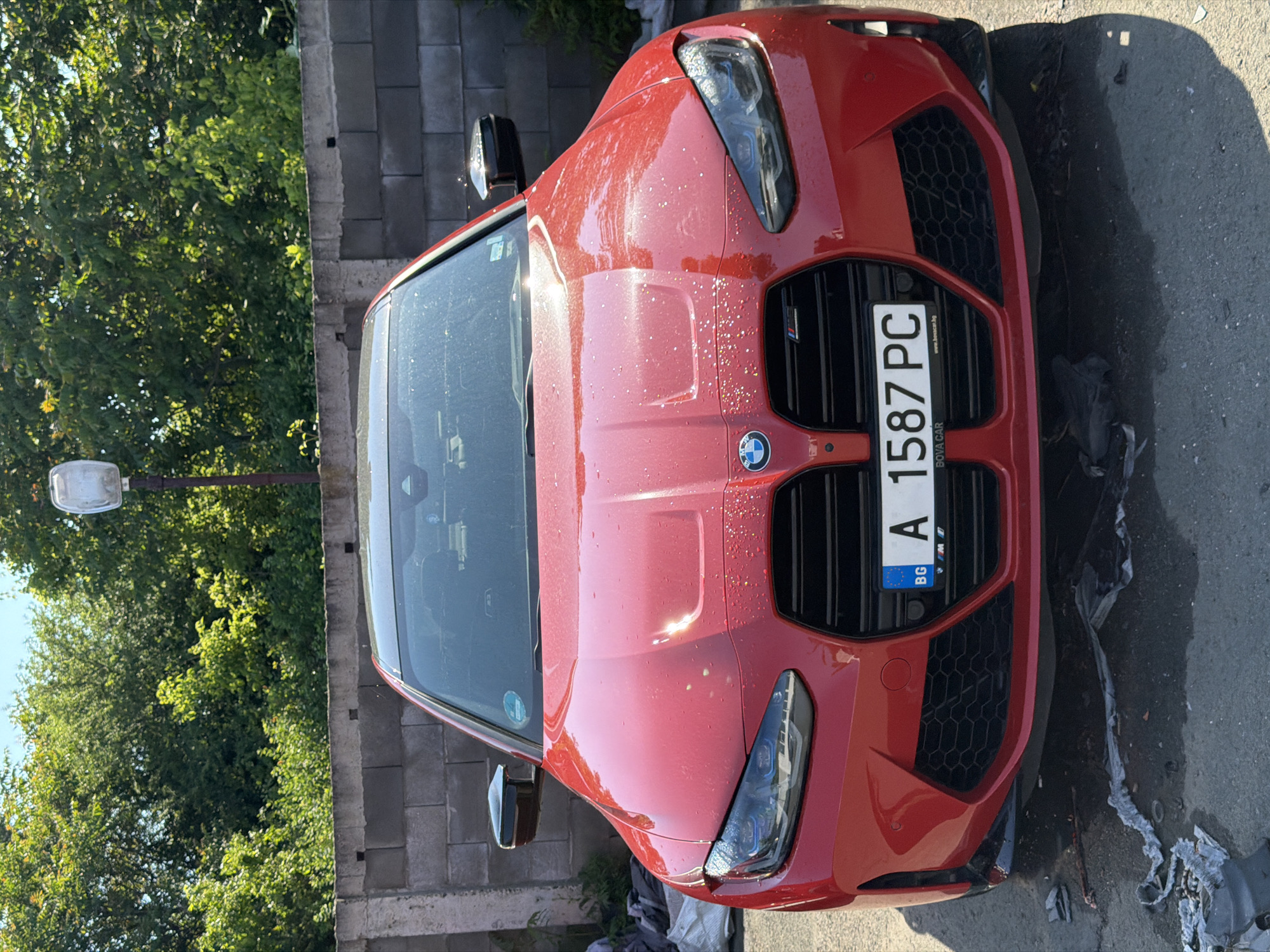The image presents a bright red BMW performance car, likely an X3 M or X4 M model, parked outdoors during a sunny day. The car is captured from a high-angle, slightly off-center perspective, showcasing its front end and hood, which are covered in numerous water droplets, suggesting it has recently been washed or caught in a rain shower.
A prominent black kidney grille, characteristic of BMW's M series, features a distinctive "M" badge on the left side and the circular BMW logo in the center. The modern, angular headlights are also visible. A Bulgarian license plate, displaying "BG" and the registration "A 1587 PC", is attached to the front grille. The "A" code indicates the car is registered in the Burgas region. The license plate holder also bears the text "BOVA CAR" and "www.bovacar.bg".
In the background to the left, dense green foliage and trees are brightly lit by the sun, casting strong shadows that suggest mid-morning or afternoon. A light pole with a street light fixture is partially visible on the far left. To the right of the car, a sturdy concrete block wall provides a contrasting backdrop. The car is parked on an asphalt surface, and some crumpled grey and black plastic debris is scattered on the ground near its front right side. The overall scene depicts a parked vehicle in an outdoor setting in Burgas, Bulgaria.