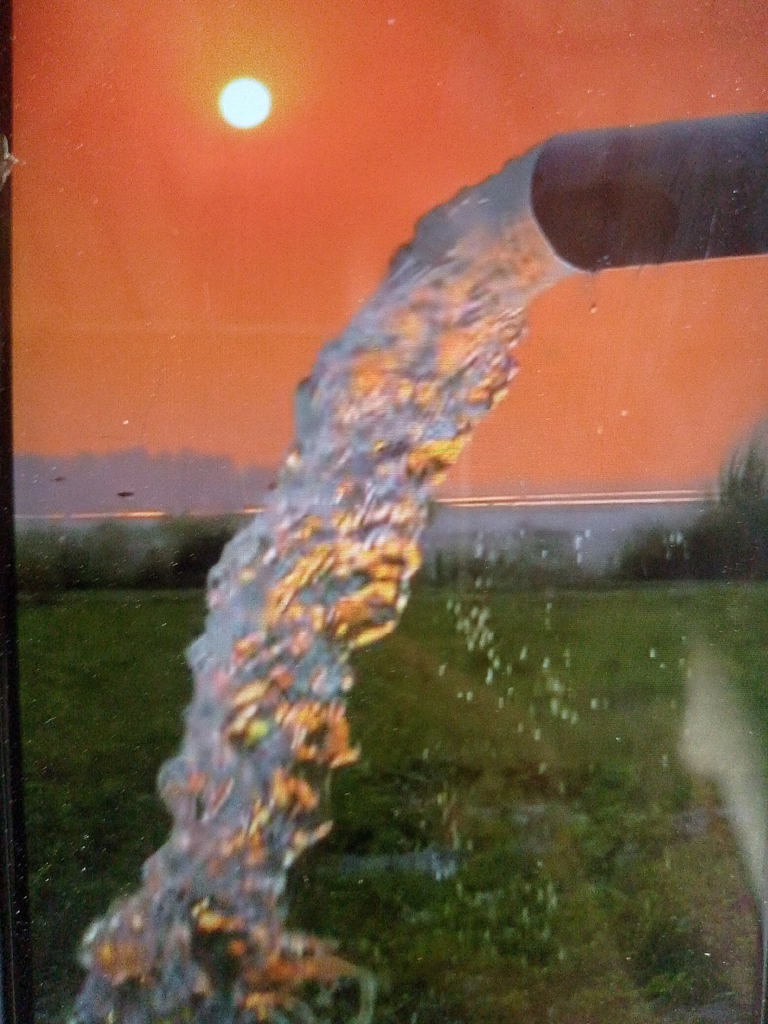 A photograph depicts water pouring from a dark-colored pipe against a backdrop of a reddish-orange sky with a bright white sun. Below the horizon, there is a blurred landscape with green vegetation. Some water droplets are visible in the foreground.