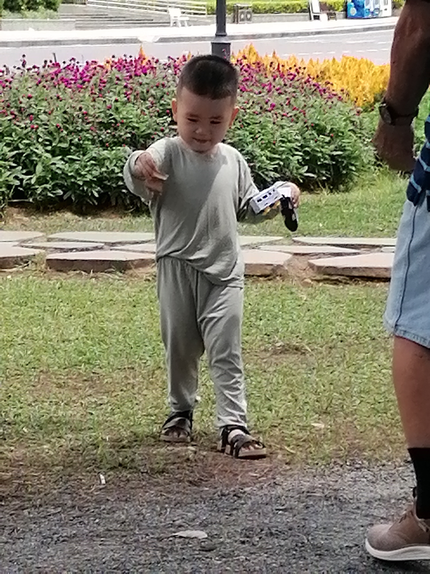 A young East Asian boy stands in a park-like setting, holding a small white toy. He is wearing a light grey long-sleeved shirt and matching pants, along with dark sandals. The boy is looking down at the toy in his hands. In the background, there are beds of pink and yellow flowers, lush green bushes, and a paved walkway. The ground is covered with grass and some gravel. To the right of the frame, the legs of an adult are visible, wearing a blue and white striped shirt and denim shorts, with a watch on their wrist. The scene appears to be a sunny day. The location is likely Da Lat, Vietnam, given the typical landscape and floral displays often found in the city.
