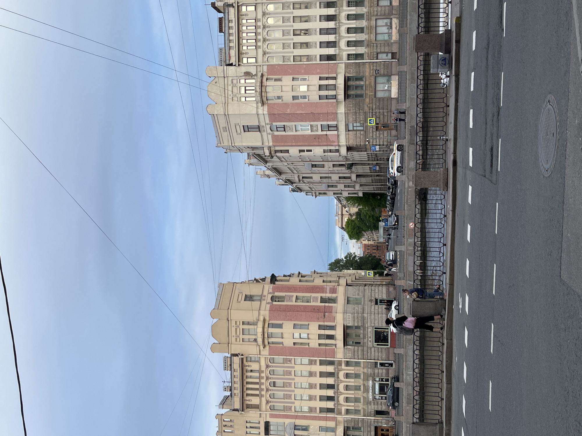 The image captures a broad street or embankment in Saint Petersburg, Russia, under an overcast or soft daylight sky. The scene is dominated by two grand, multi-story buildings flanking what appears to be a canal or river, though the water itself is not fully visible.
The buildings exhibit characteristic Saint Petersburg architecture, featuring ornate facades with decorative elements, arched windows, and varying textures. They are constructed with a mix of light beige or cream-colored stone and sections of pinkish-red brickwork. The building on the left side of the frame (when viewed correctly) appears to have a more intricate, stepped roofline.
In the foreground, a paved road with white dashed lane markings stretches into the distance. A sturdy metal railing with stone posts separates the street from a sidewalk and the unseen water body. Several vehicles are parked along the far side of the road, and others are visible further down the street. Overhead wires crisscross the sky, suggesting the presence of public transport like trams or trolleybuses.
Several individuals are present, mostly on the sidewalks or leaning against the railing, observing the surroundings. On the right, two people are near the railing, while on the left, a person with a pink backpack stands by the embankment.
Notable details include street signage: a yellow triangular sign indicating a pedestrian crossing and blue 'P' signs for parking. A prominent blue sign on the railing clearly states "НЕ БРОСАТЬ МУСОР В ВОДУ" (Do not throw trash into the water). Other visible text on storefronts includes "ШИНОМОНТАЖ" (Tire fitting) and "ЦВЕТЫ ПОДАРКИ" (Flowers Gifts) on the right building, and "РЕМОНТ" (Repair) on the left. The light suggests it's daytime, possibly late afternoon or an overcast day, lending a serene atmosphere to the urban landscape.