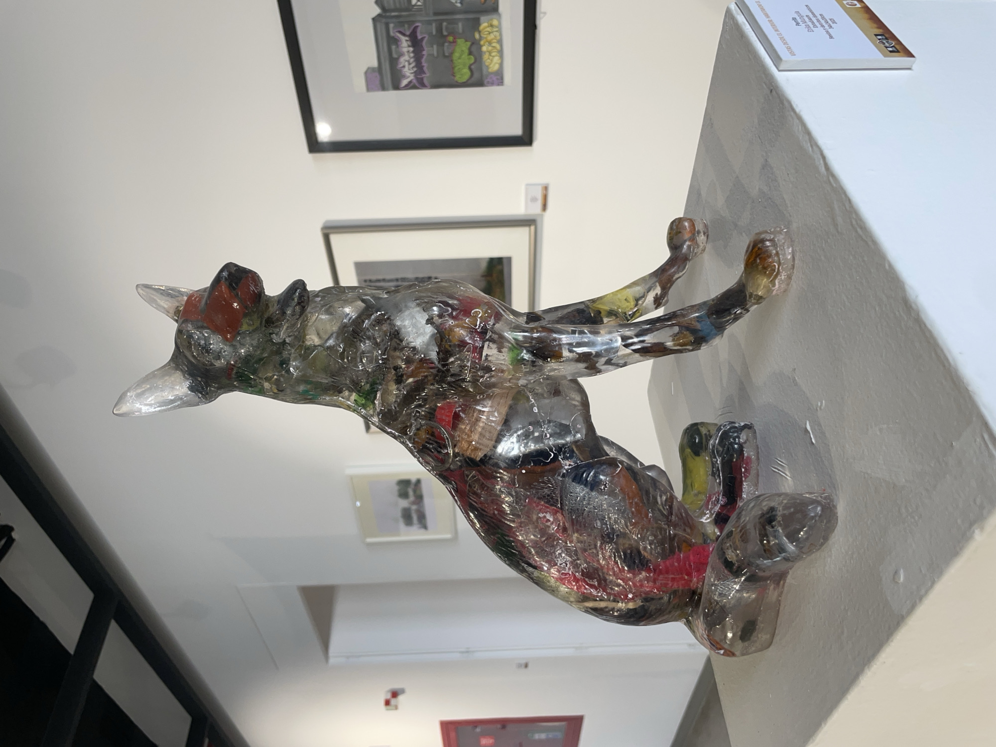 An indoor scene within an art gallery in San Andrés Cholula, Mexico, displays a transparent sculpture of a seated dog, resembling a German Shepherd, positioned on a rectangular white plinth. The sculpture is crafted from clear resin, encasing various colorful discarded materials, predominantly identifiable as plastic waste and fabric scraps. The dog's muzzle and nose area are distinctively colored in a reddish-orange hue.
The background consists of plain white walls, a visible black ceiling beam system, and several framed artworks. To the left, a framed image depicts a natural landscape. To the right, another framed artwork illustrates stylized urban buildings with graffiti-like elements in purple, green, and yellow. A small exhibition plaque is affixed to the lower right side of the white plinth. The plaque text, in Spanish, identifies the artwork as "Perrito" (Little Dog) by artist Erika Munguía, created from "Resina y diversos desechos domésticos" (Resin and various household waste) in 2023. No individuals are visible in the scene. The lighting is uniform, suggesting artificial illumination typical of a gallery setting.