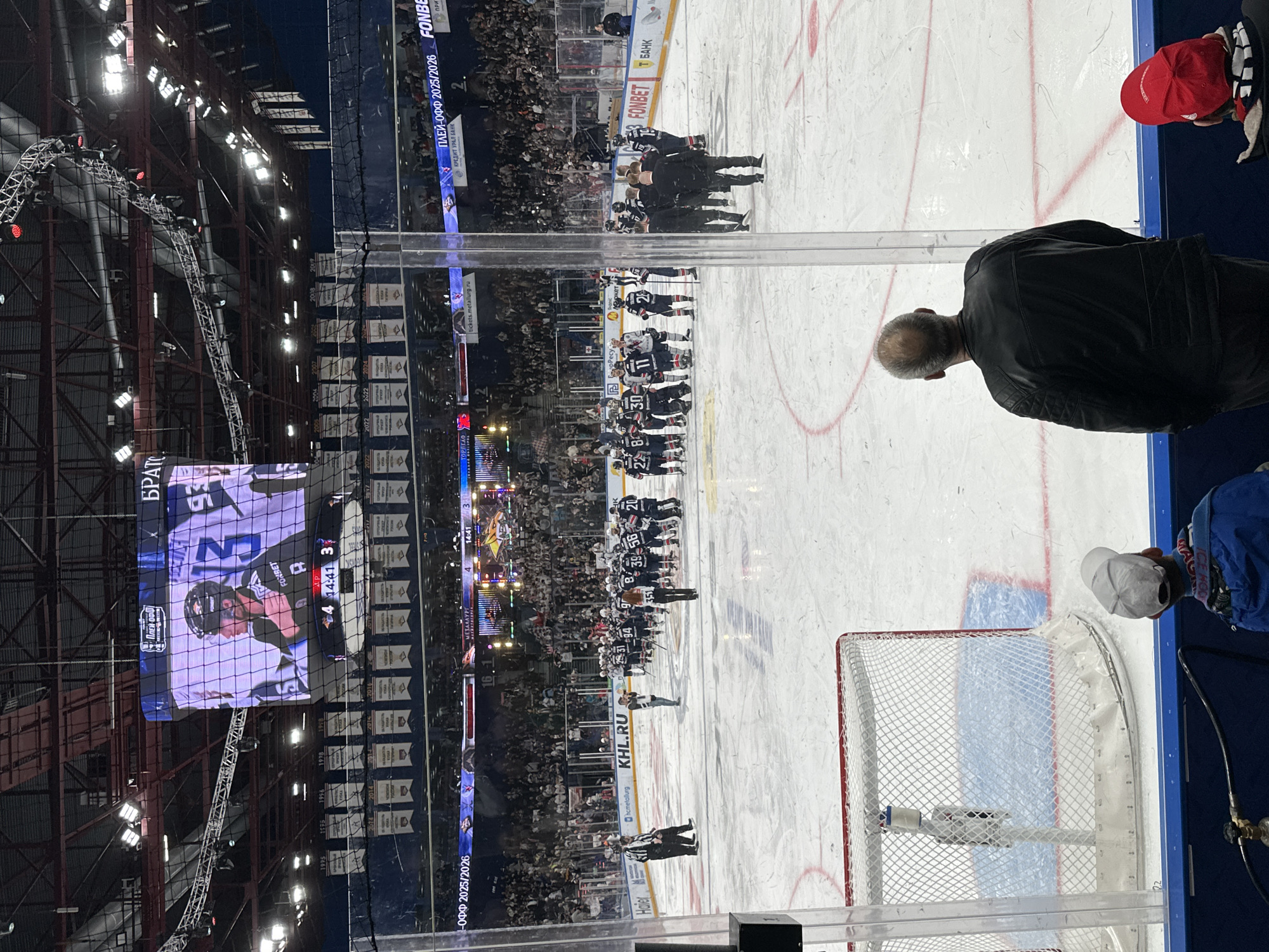 The image shows a hockey game in progress at what appears to be an indoor arena in an unknown city. The two teams, one in dark jerseys and the other in white, are lined up on the ice facing each other. Several referees are present on the ice near the teams. A large video screen shows a close-up of a player's face and game information, including the score and time remaining (4-3, 14:41). Banners with team names and logos hang on the side walls of the arena, and a large crowd of spectators fills the stands. In the foreground, people are observing the game from behind a glass barrier, one is wearing a black leather jacket. Part of a hockey net can be seen.
