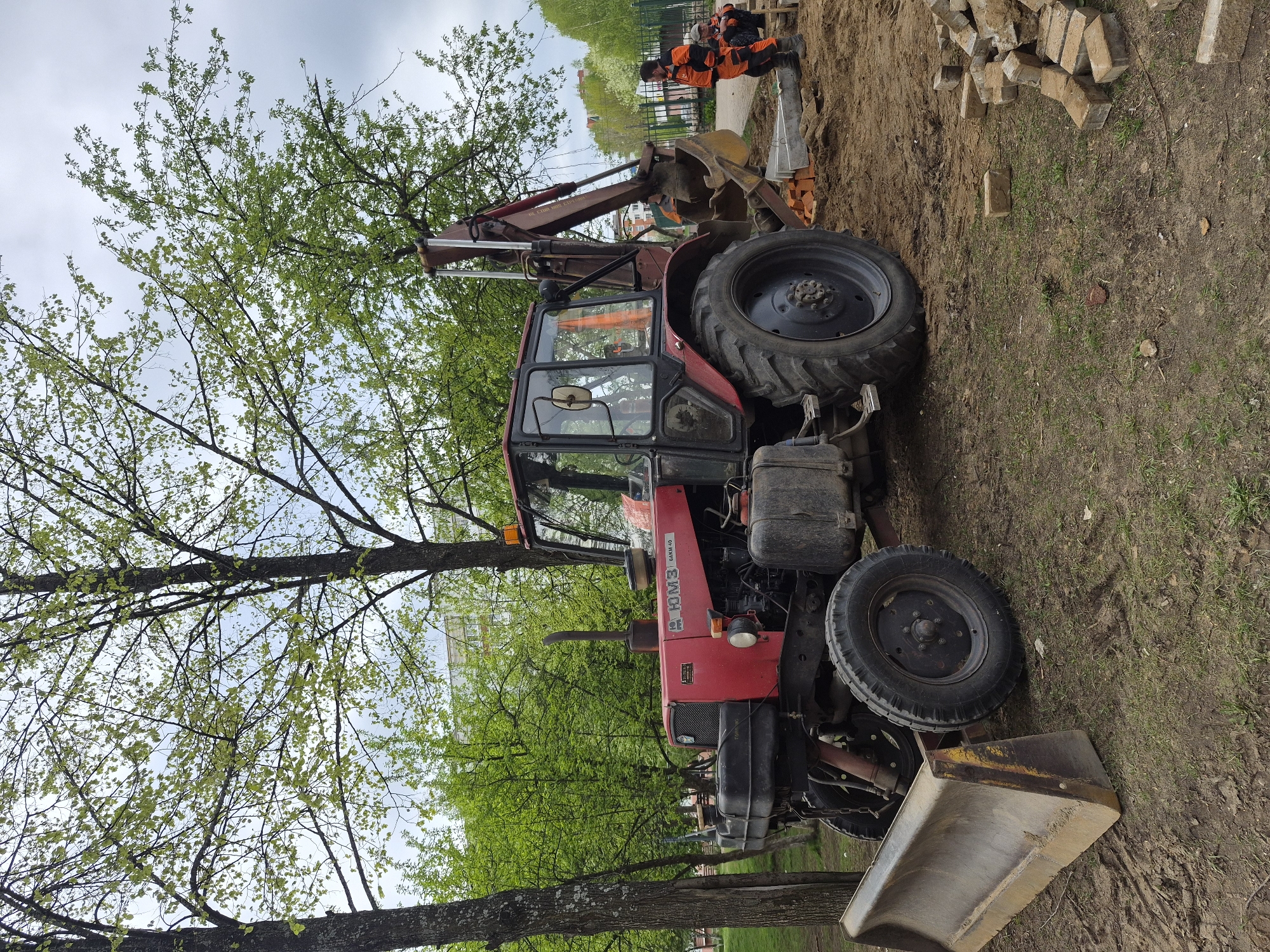 A red tractor with a front-end loader and backhoe attachment is parked on a dirt and grass area next to a sidewalk in an urban setting in Unknown, Unknown. The tractor is facing right, and its front-end loader bucket rests on the ground. The backhoe arm is raised and partially obscuring two individuals wearing orange safety vests who are working on a section of sidewalk next to a pile of paving stones. Trees with green foliage are visible throughout the scene, and a building is faintly visible in the background.